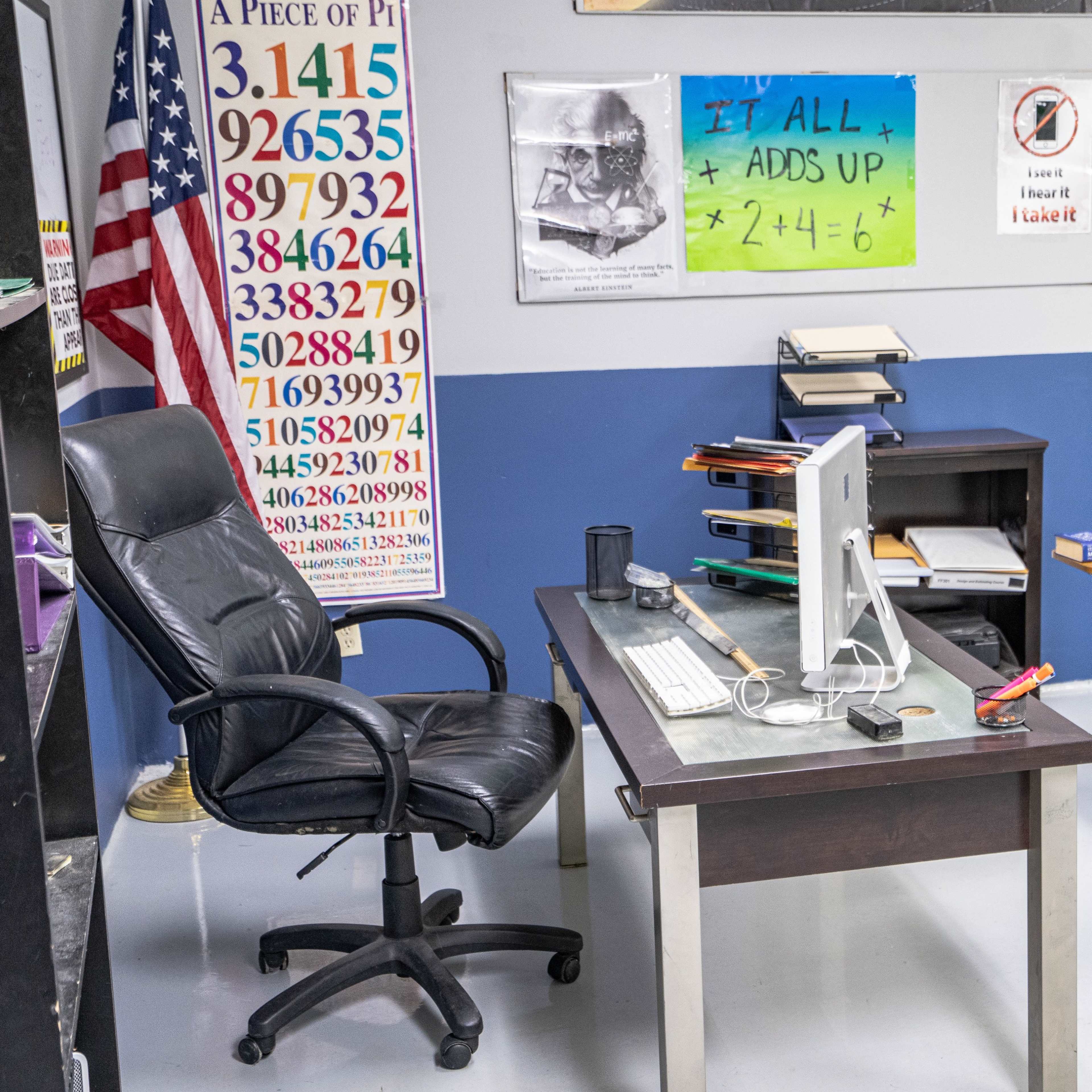 A classroom is set up with a desk and computer in front, chairs arranged for students, and educational materials displayed on the walls.