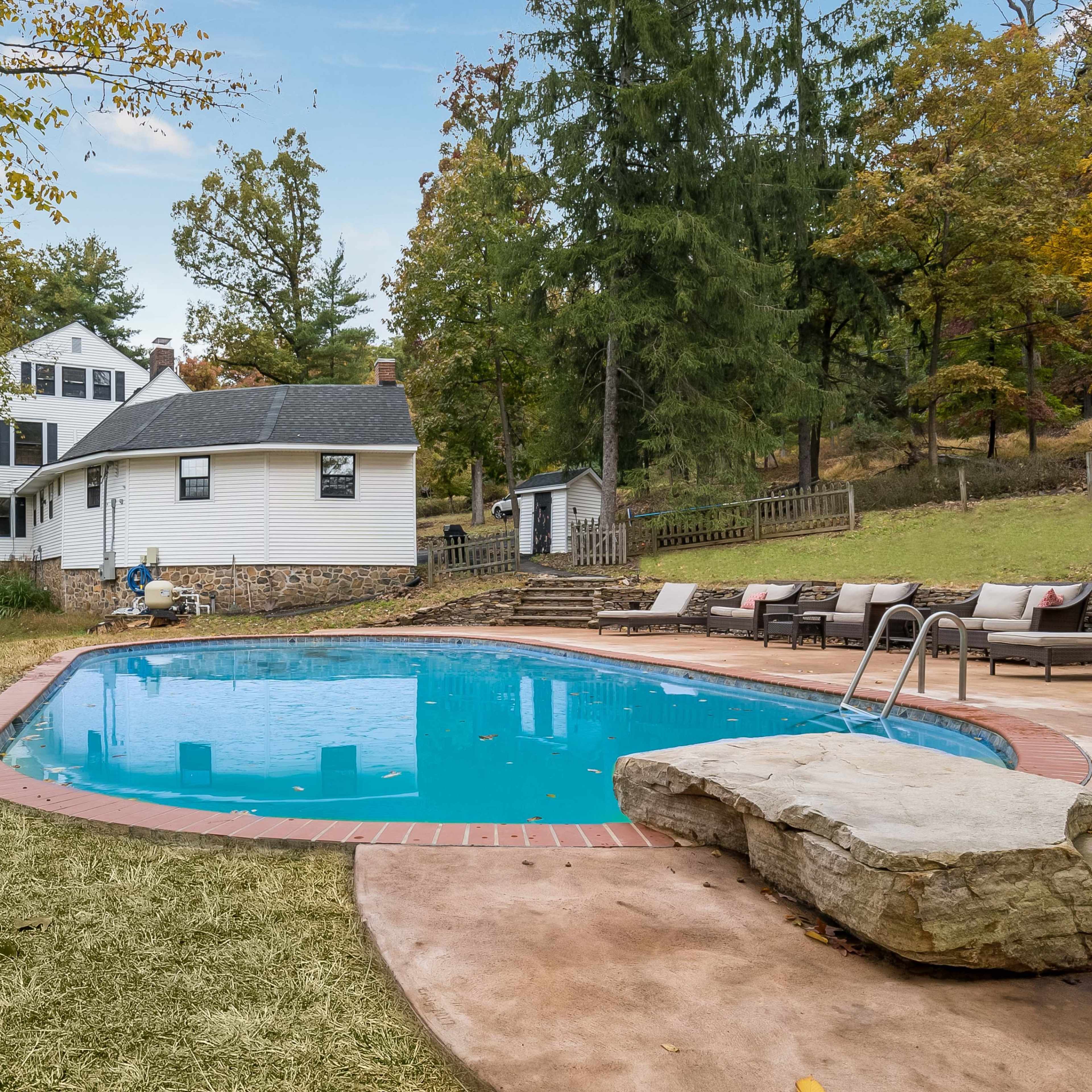 A swimming pool is situated in the backyard of a white house, surrounded by lounge chairs and trees with autumn foliage.