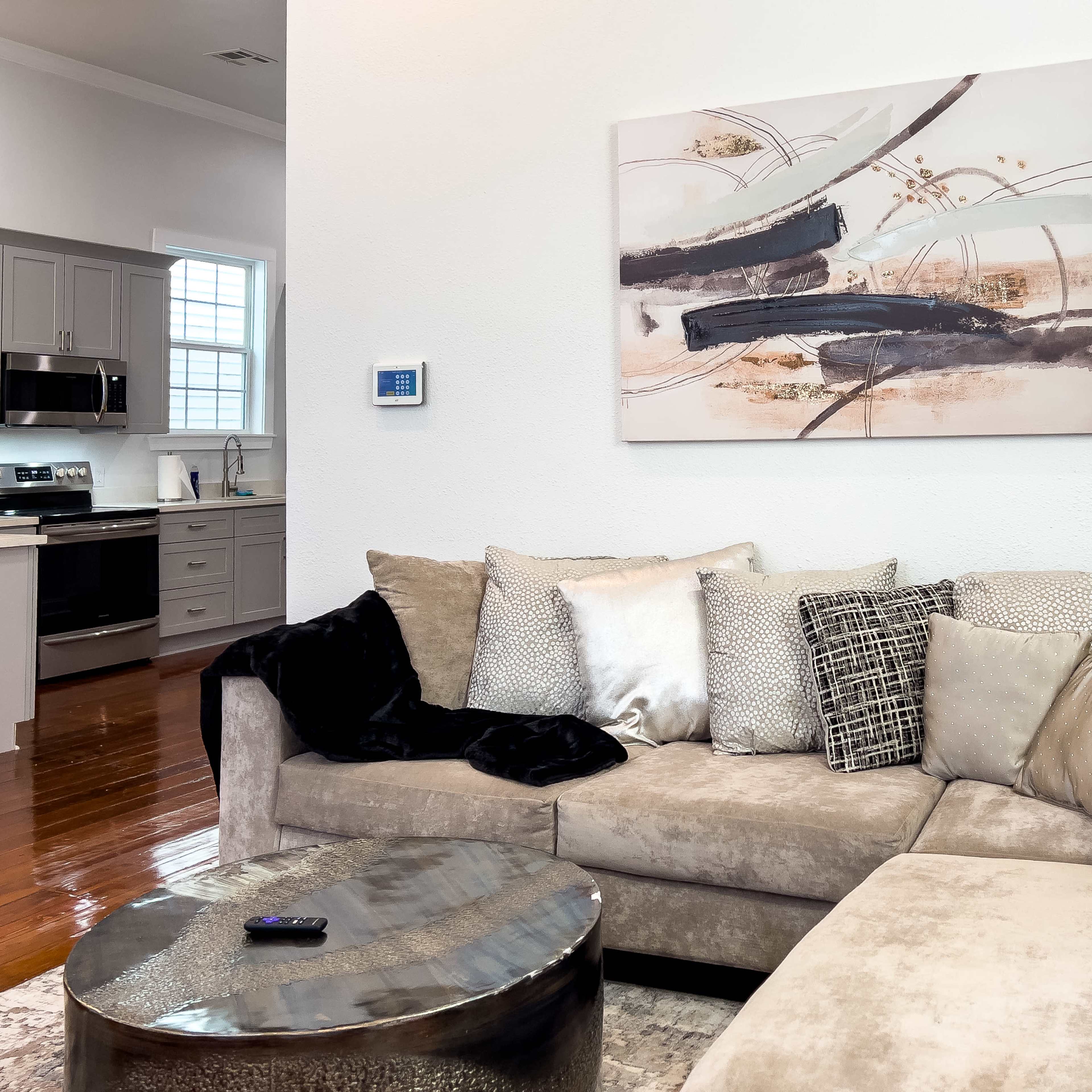 A modern living room features a sectional sofa with various pillows, a round metallic coffee table, and abstract wall art, with a glimpse of a kitchen in the background.