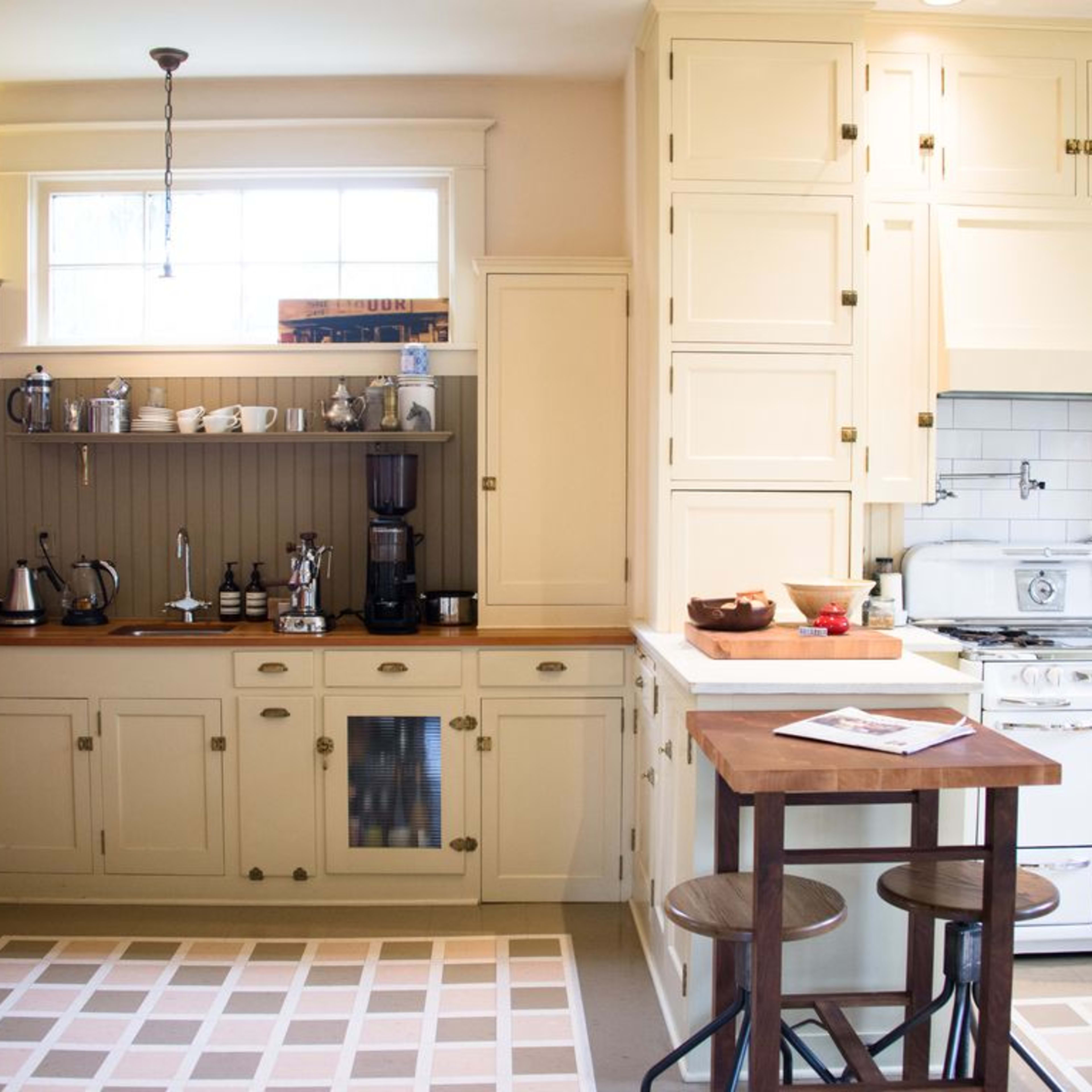 A modern kitchen featuring light-colored cabinets, a white stove, a coffee maker, and a small dining table with two stools against a backdrop of a patterned rug.