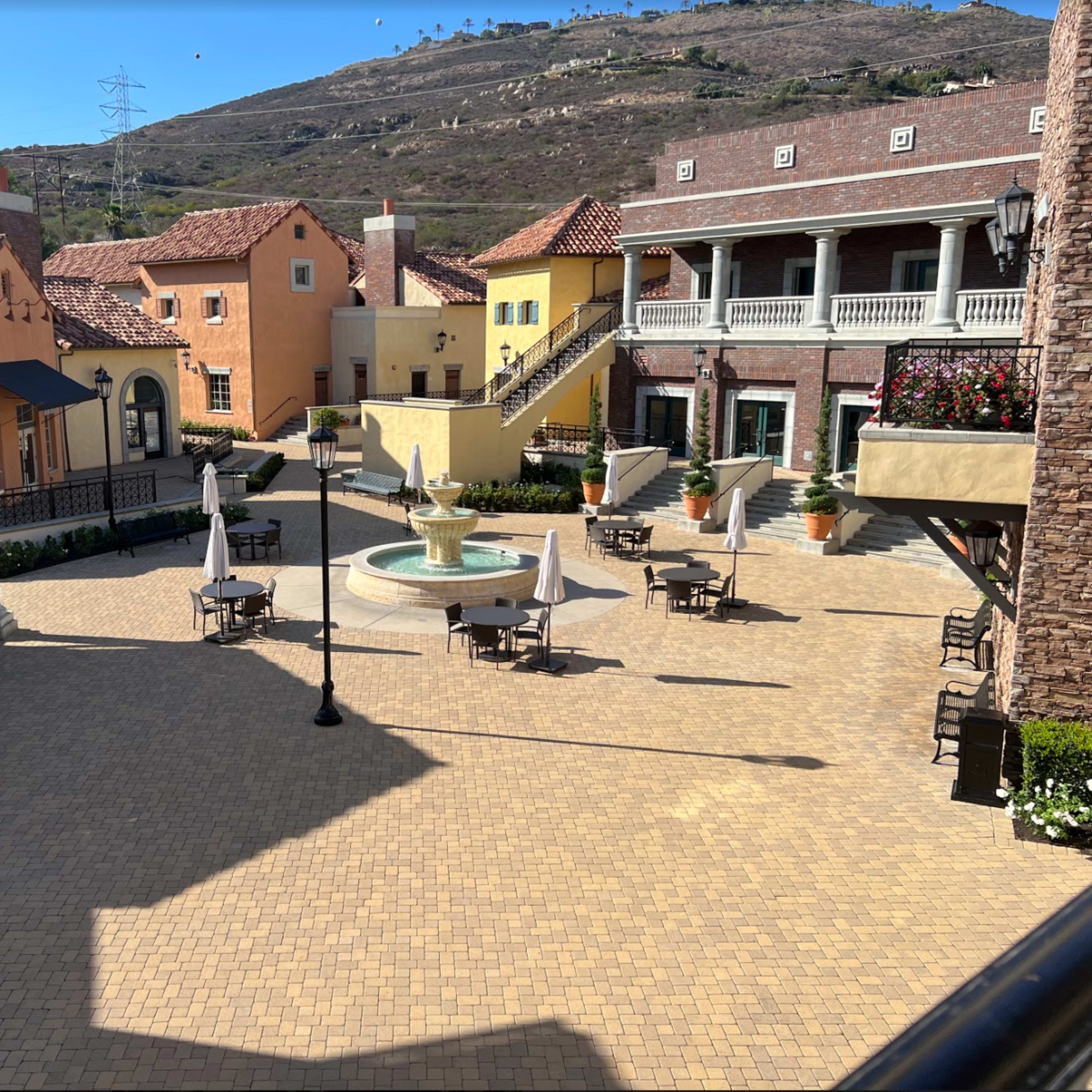 A courtyard with a fountain at the center, surrounded by several buildings featuring a mix of architectural styles and outdoor seating areas.