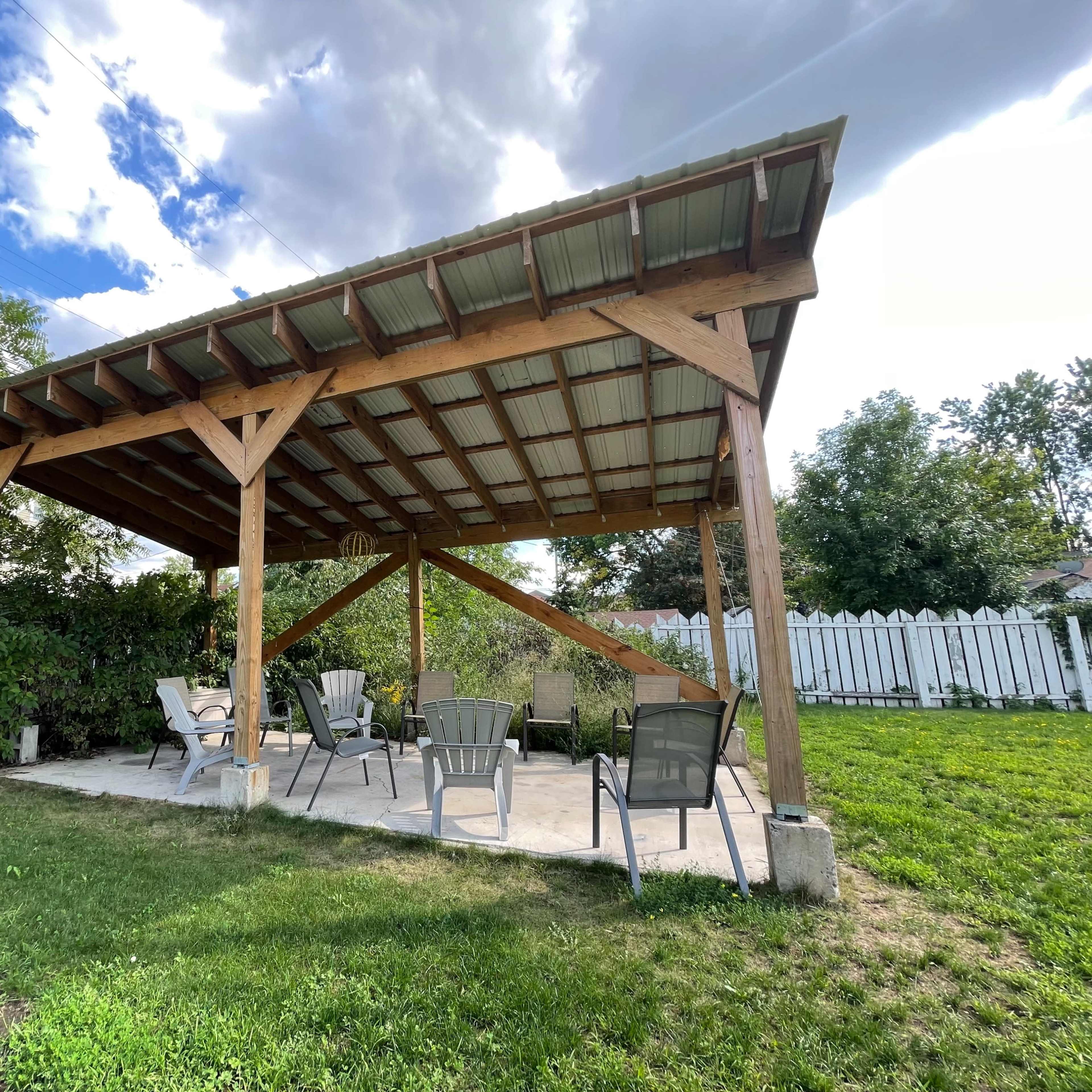 A wooden pavilion with a metal roof stands on a concrete patio, surrounded by greenery and facing a white picket fence.
