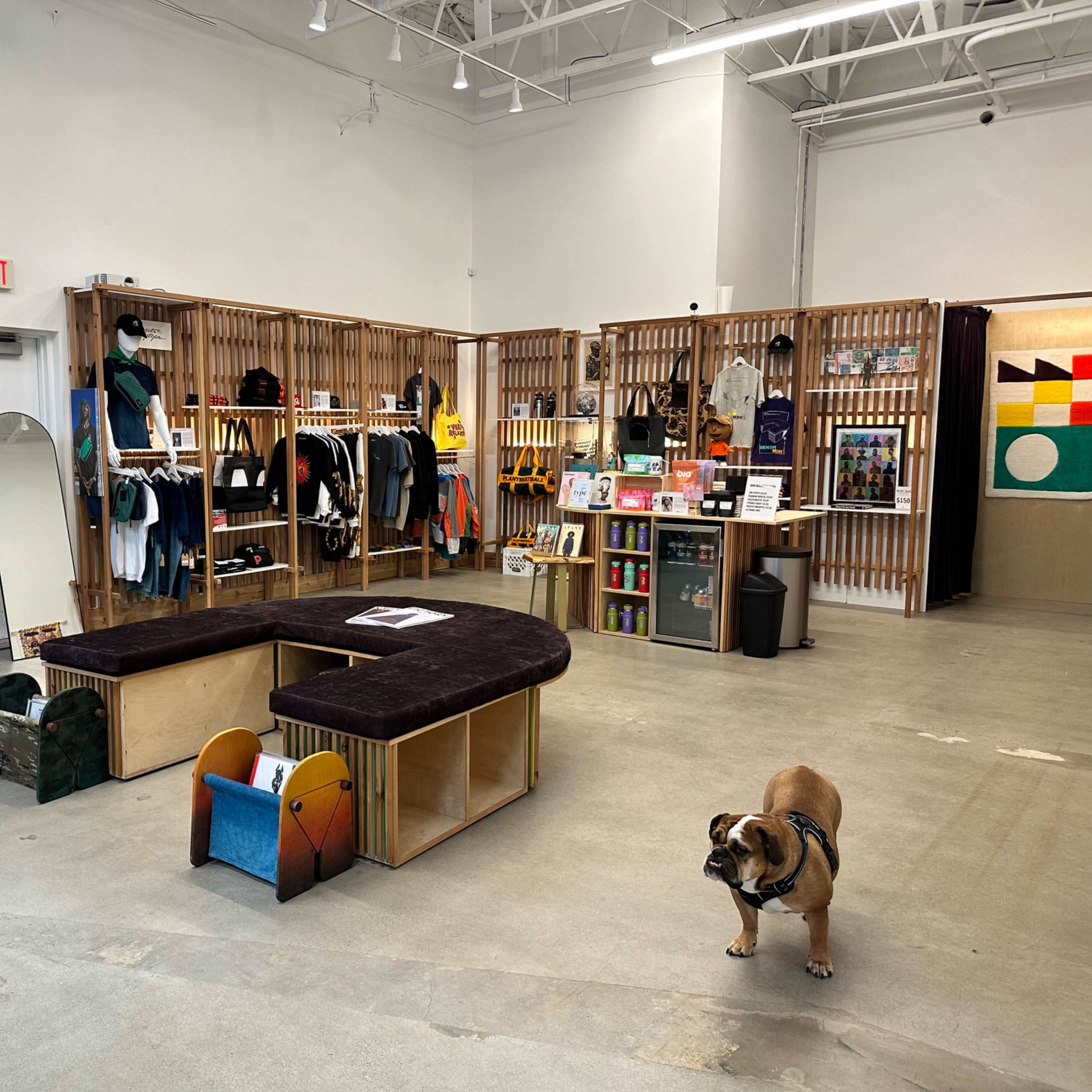 A retail store with wooden shelving displaying clothing and merchandise, a central seating area, and a dog on the floor.