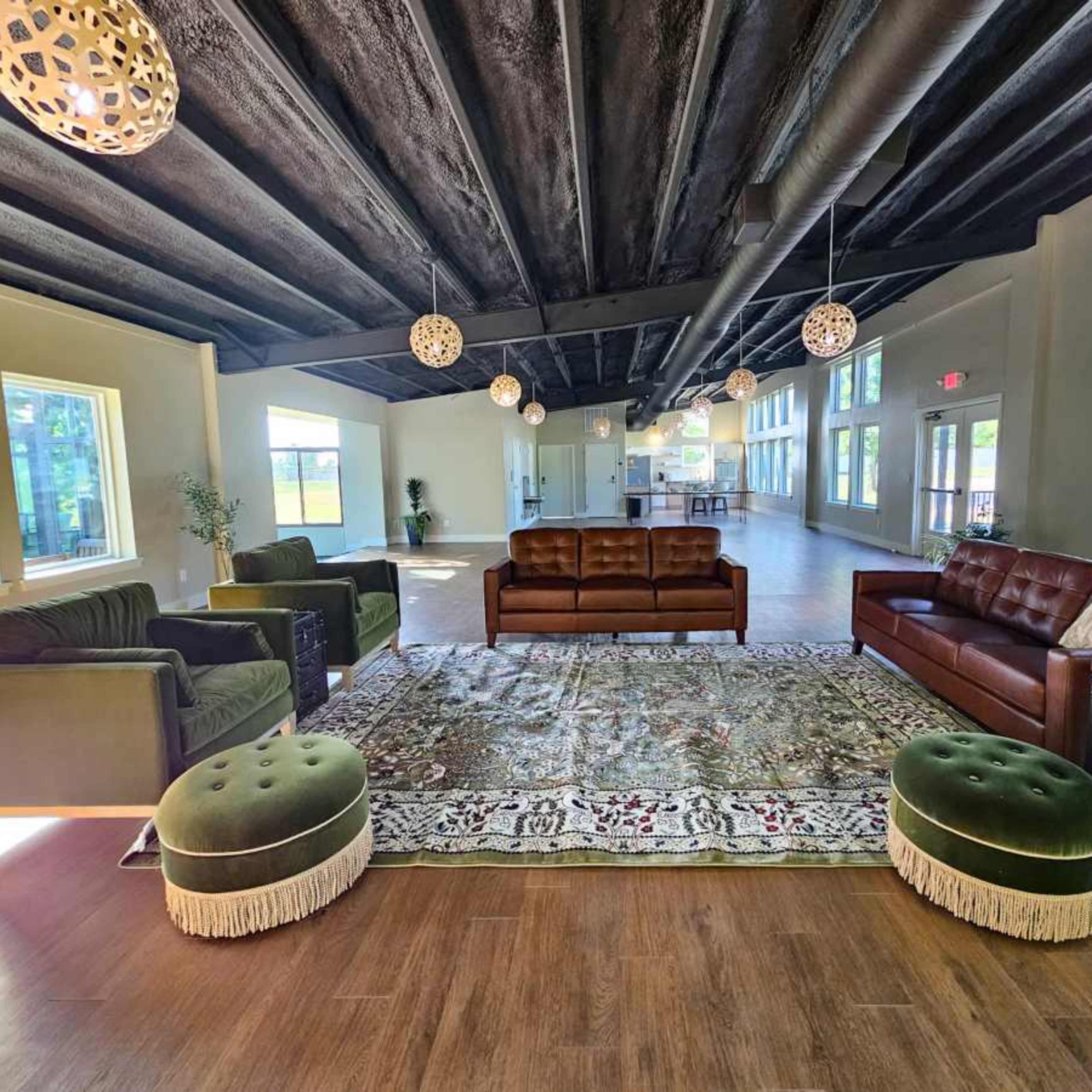 A modern lounge area with several sofas, green ottomans, and a large area rug under decorative light fixtures and spacious windows.