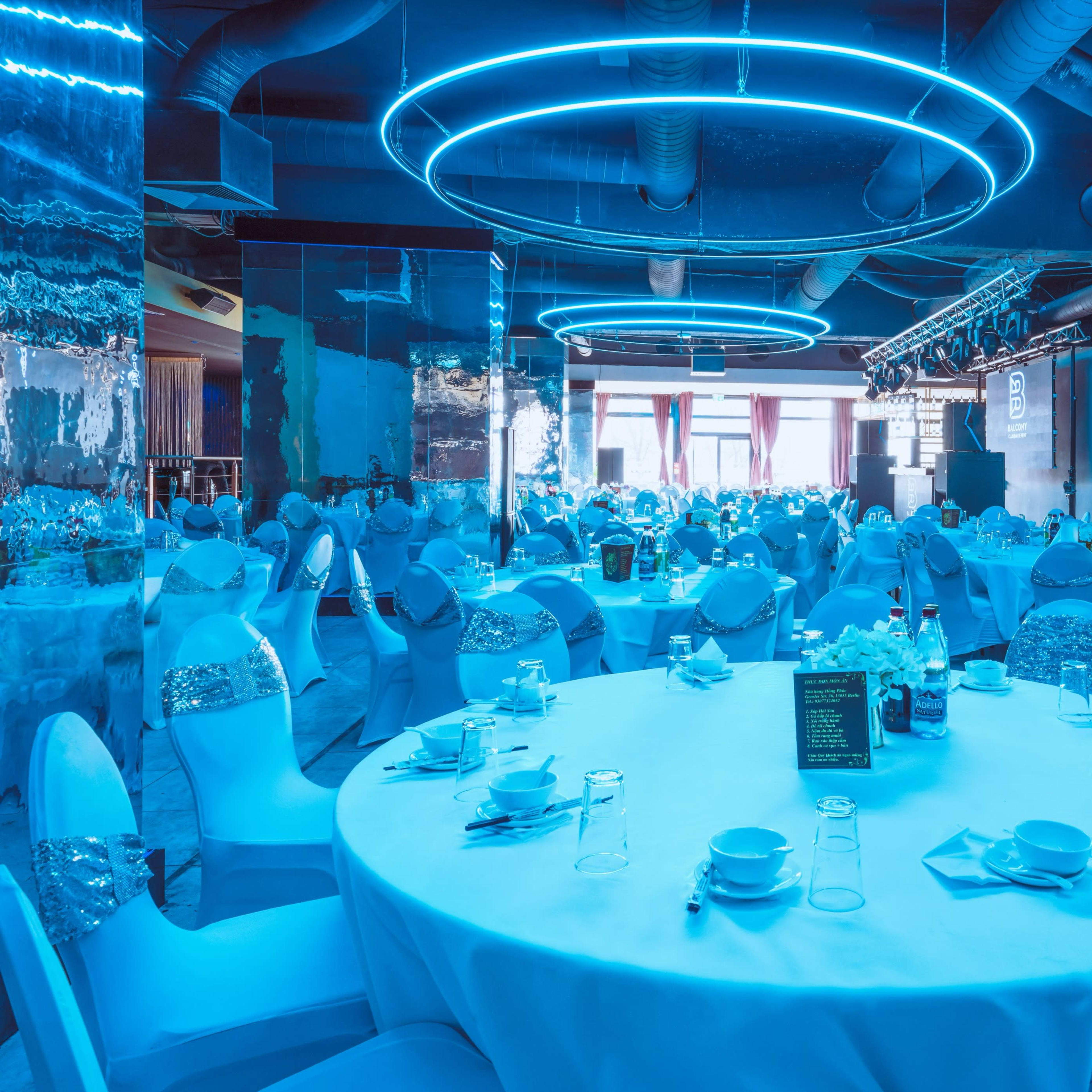 A spacious banquet hall is set up with round tables covered in white tablecloths and decorated with blue lighting.