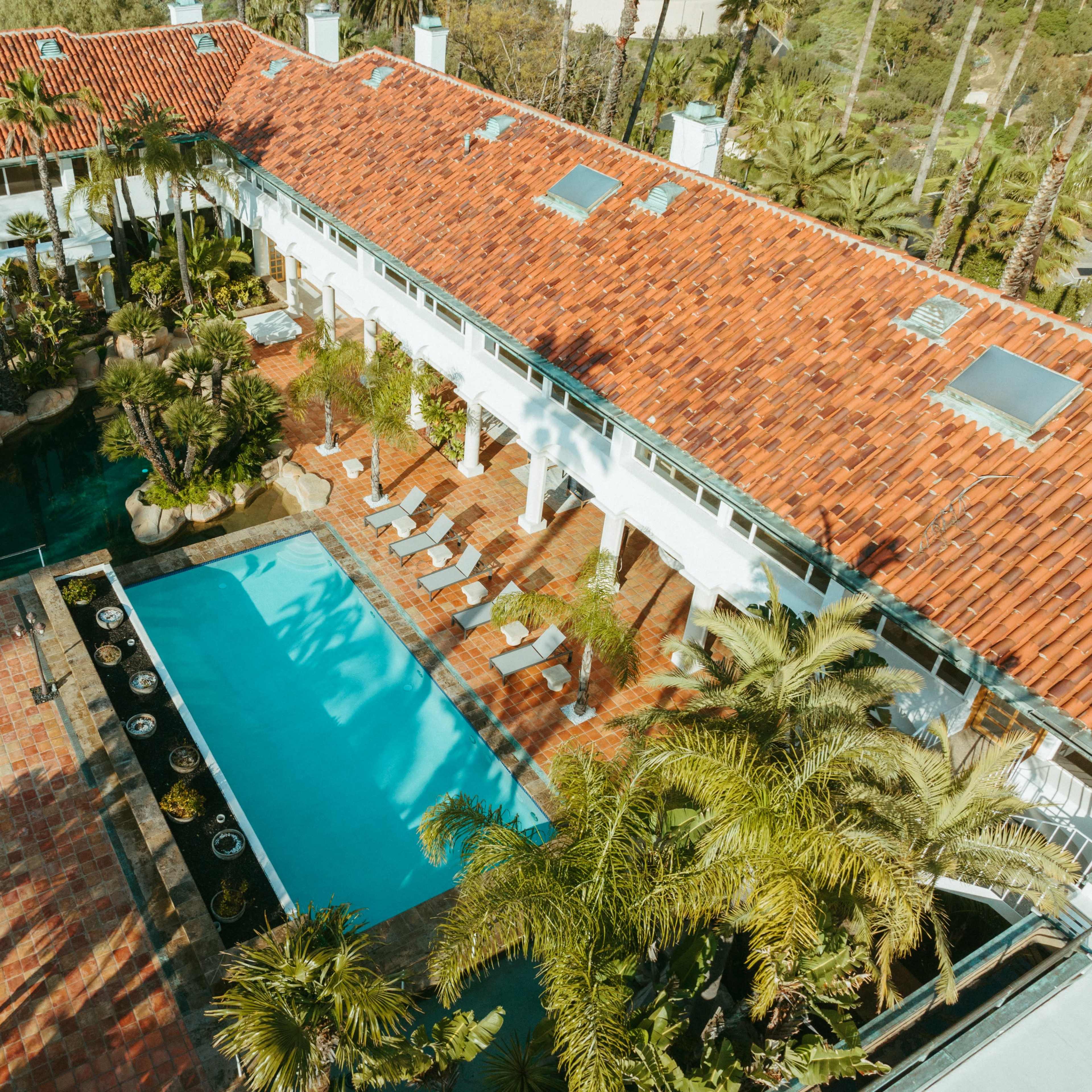 A rectangular swimming pool surrounded by palm trees and lounge chairs, adjacent to a large, red-tiled building.