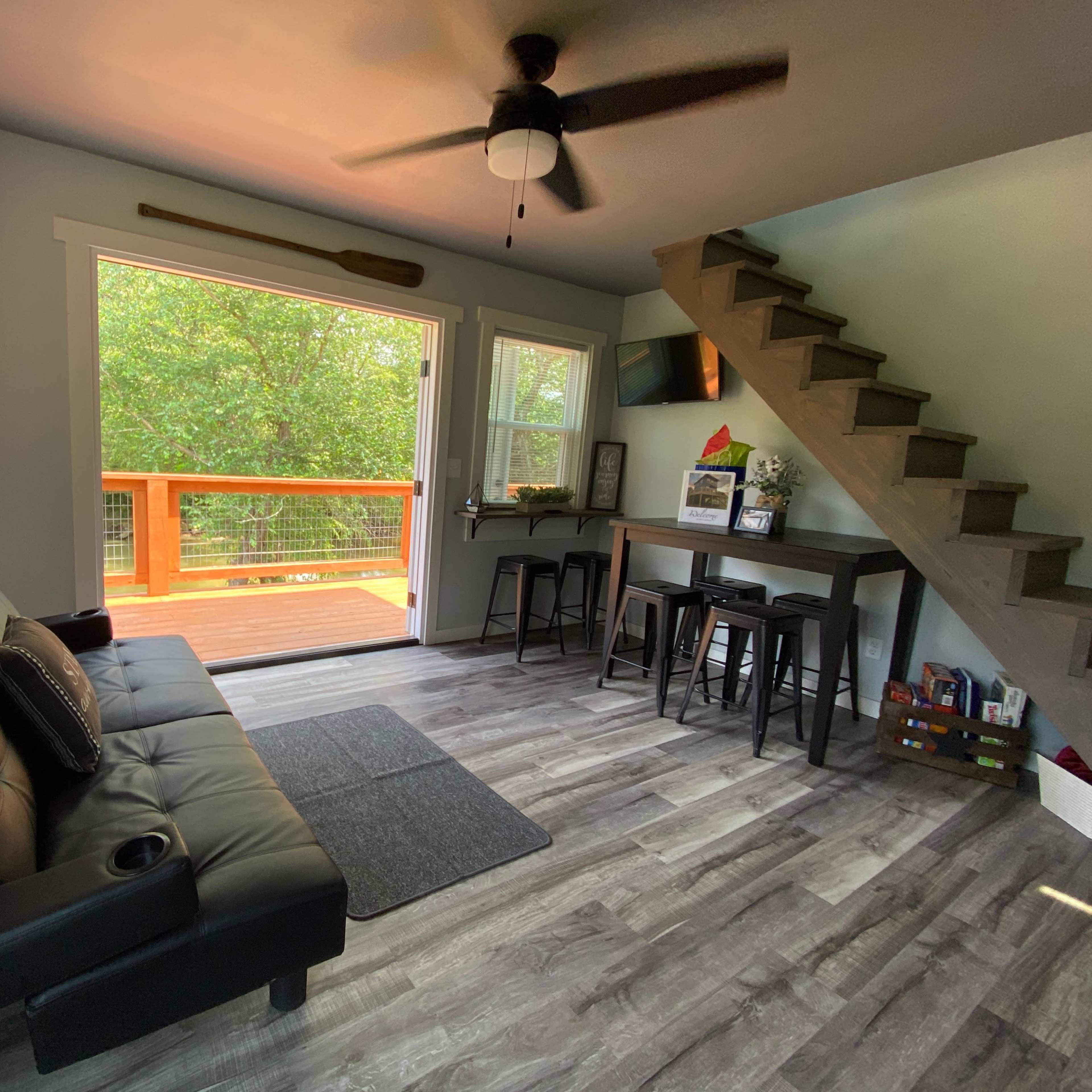 A modern living area with a black futon, a small dining table, and a staircase leading to an upper level, with large windows overlooking a deck.