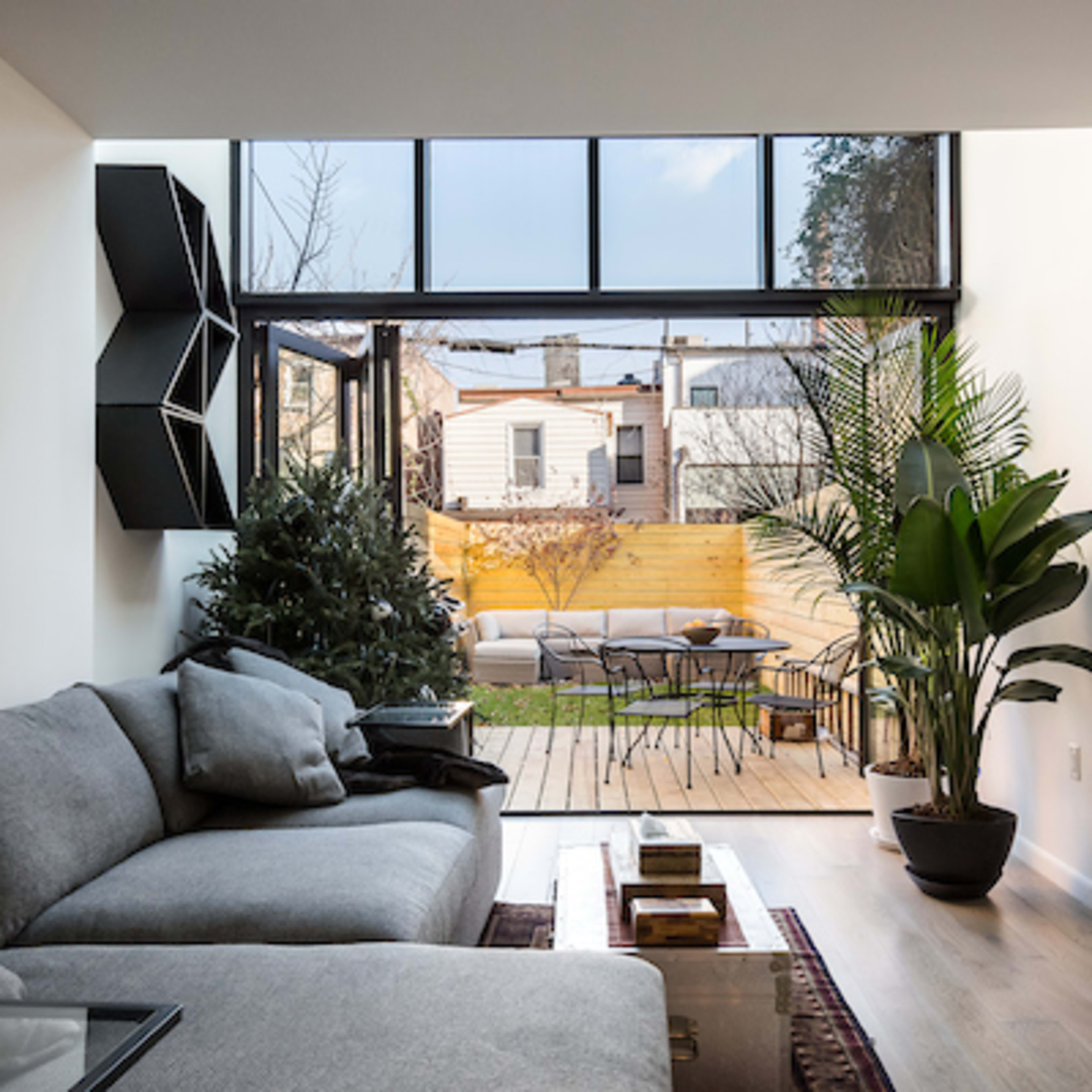 A modern living room features a gray sofa, large windows that open to a backyard, and a view of patio seating and greenery.