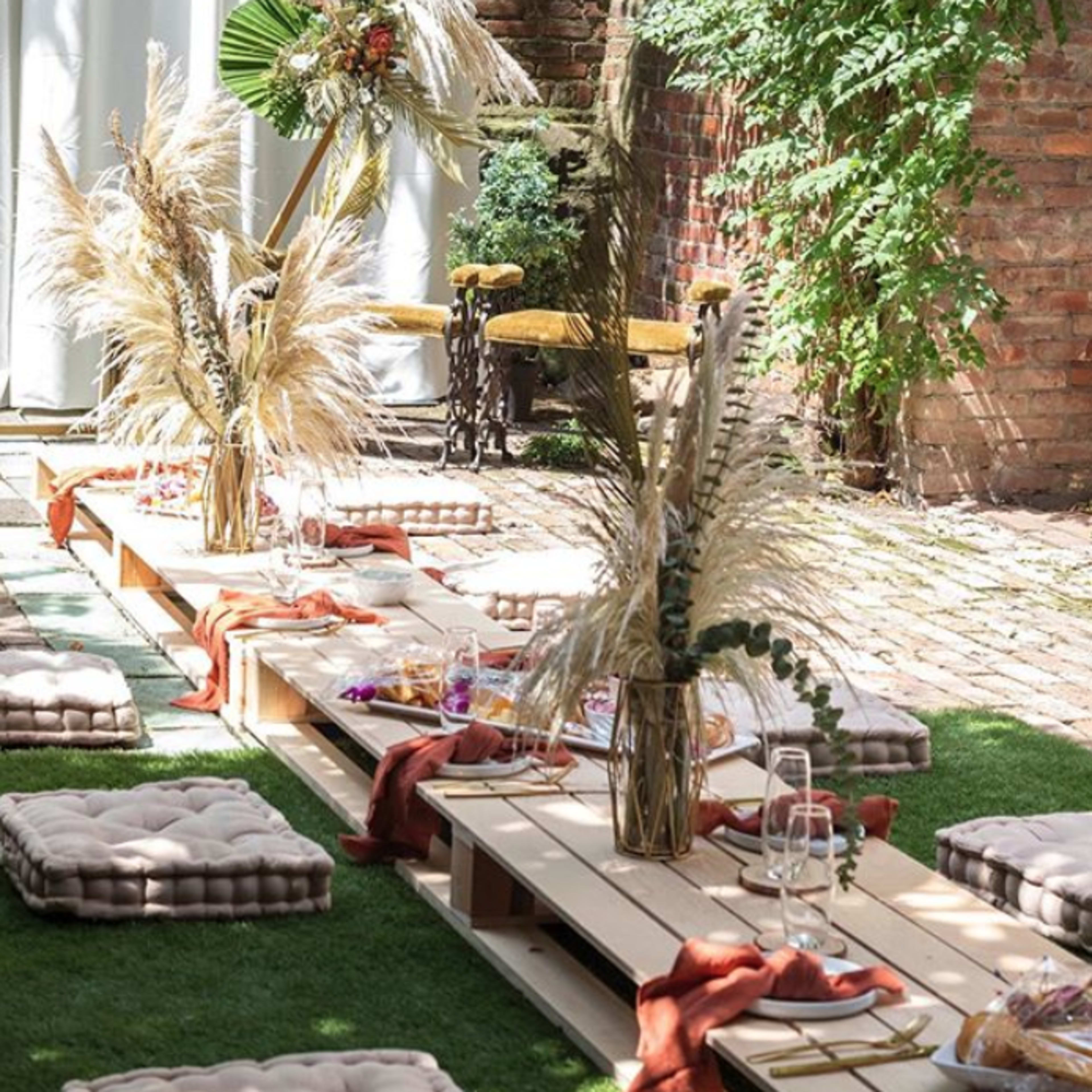 A decorated outdoor space with a wooden platform adorned with cushions and floral arrangements, surrounded by greenery and a backdrop of a white curtain.