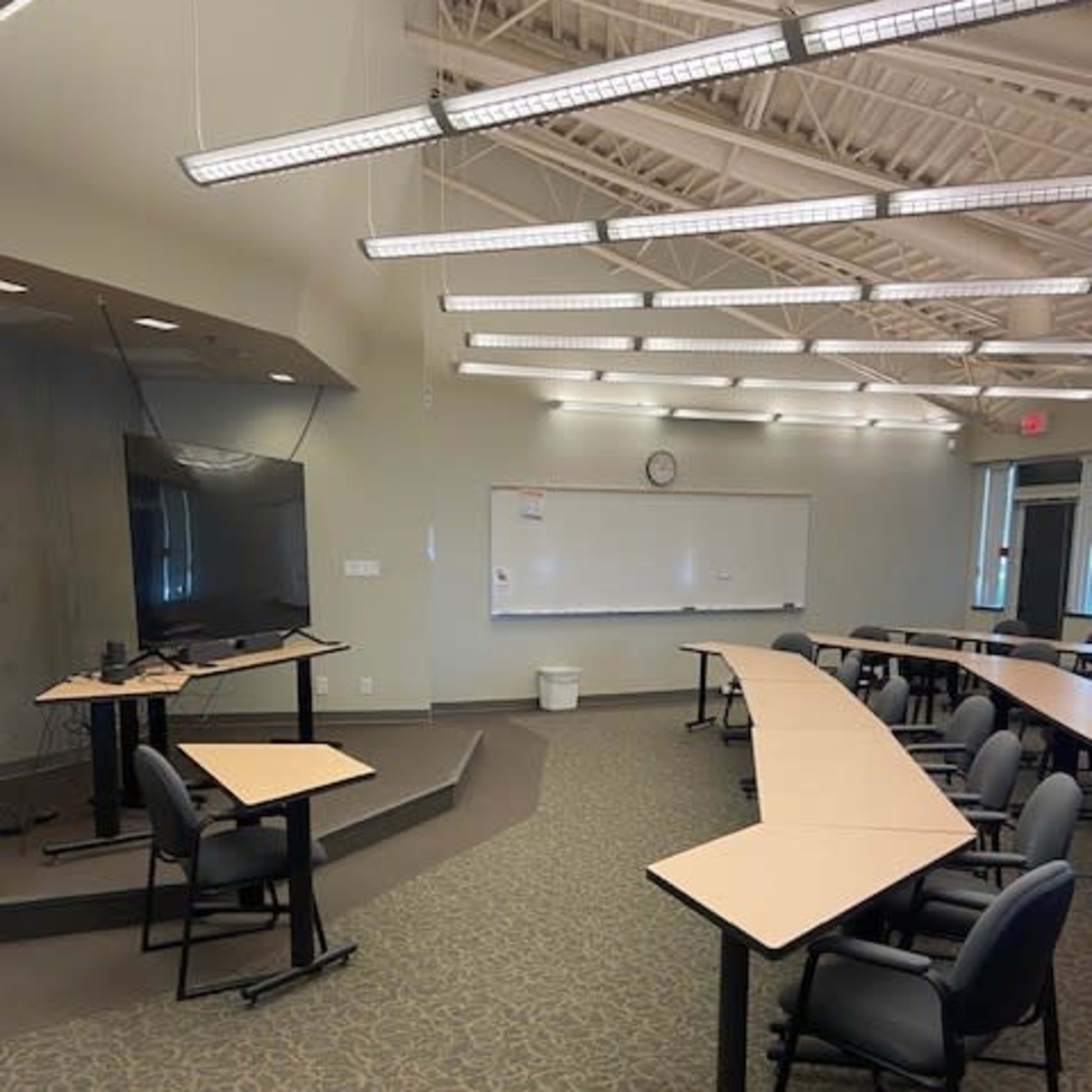 A classroom setup with curved desks and chairs, a television screen, and a whiteboard on the wall.