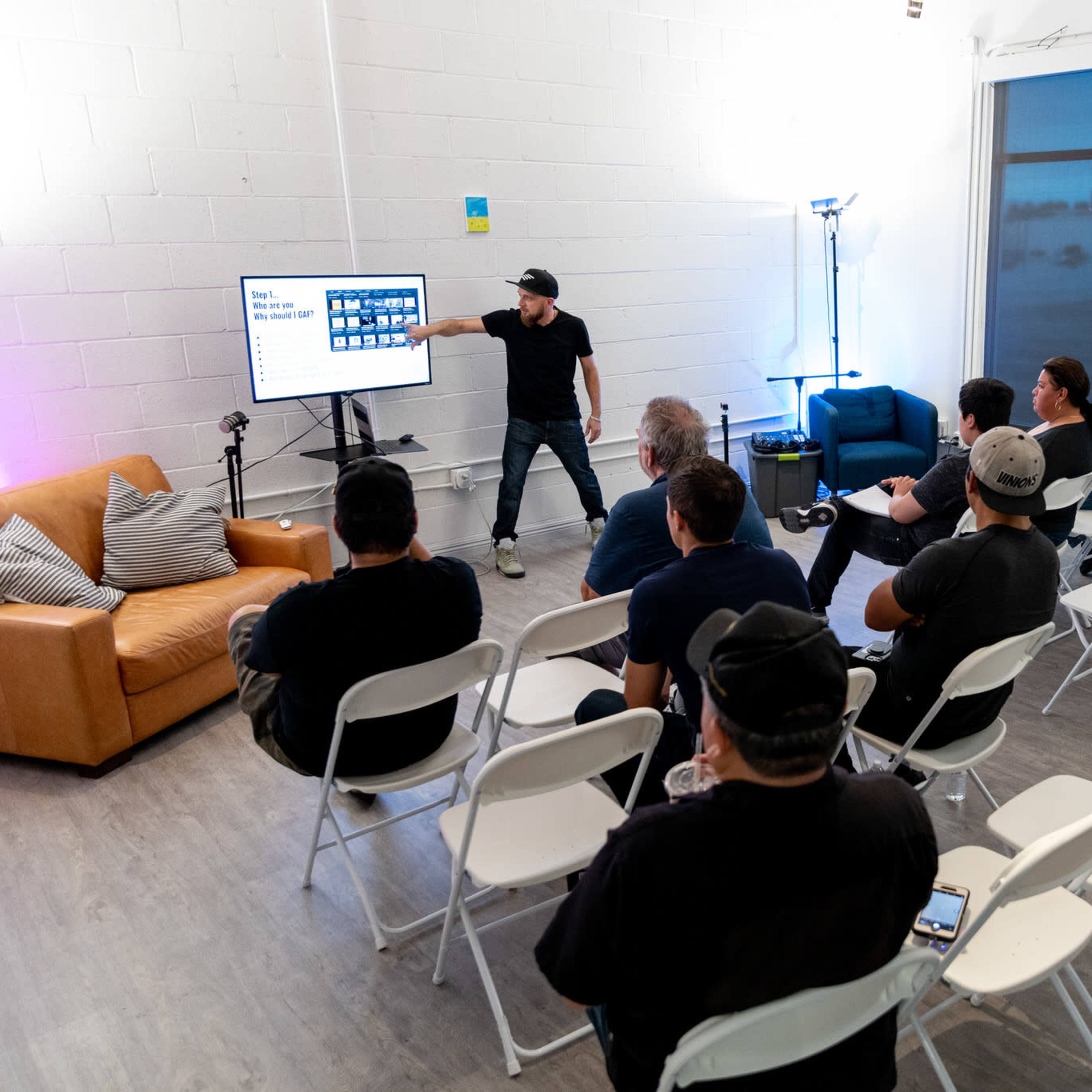 A presenter stands in front of a small audience seated in white chairs, pointing at a screen displaying various images in a well-lit room.