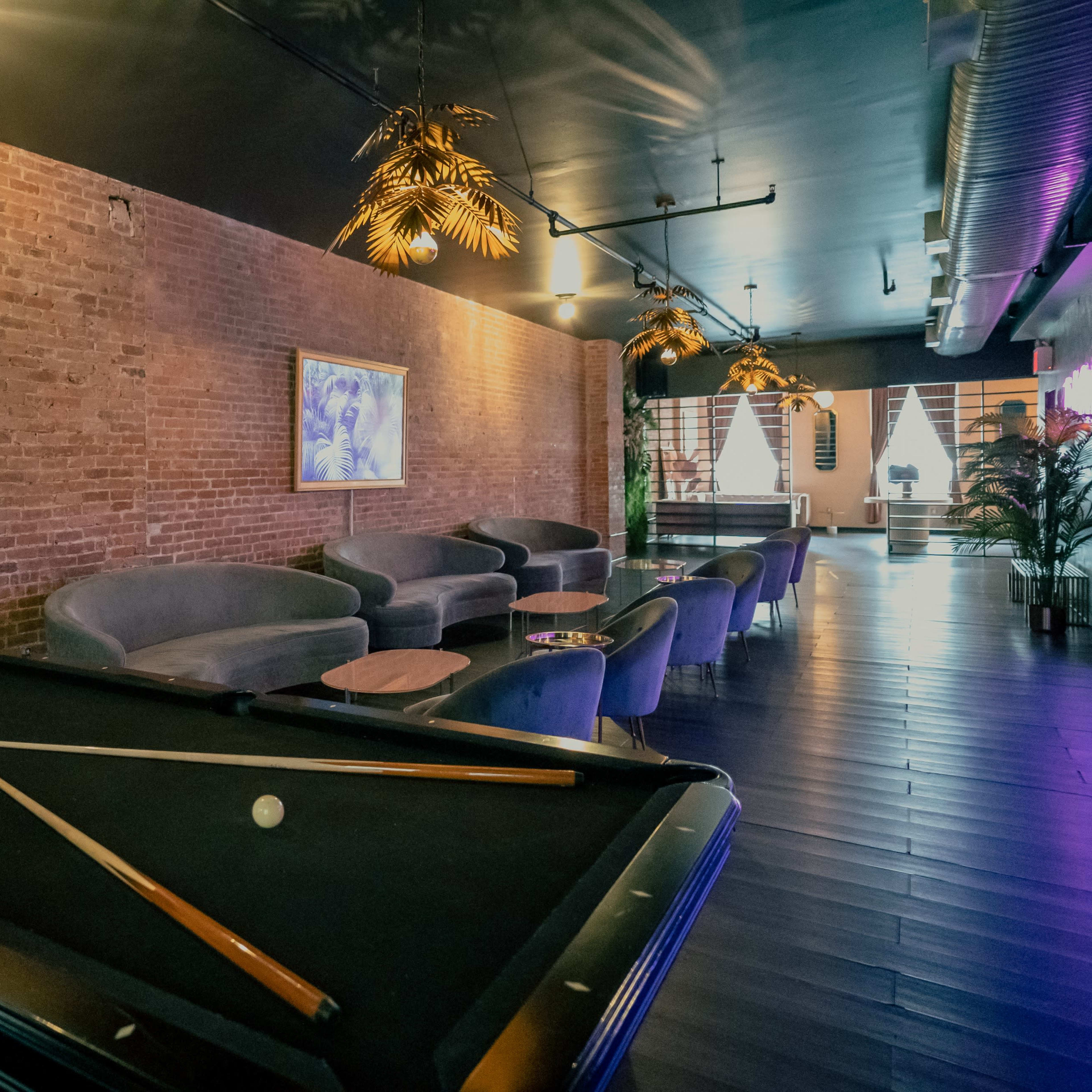 A modern lounge area featuring a billiard table, gray furnishings, exposed brick walls, and decorative lighting.