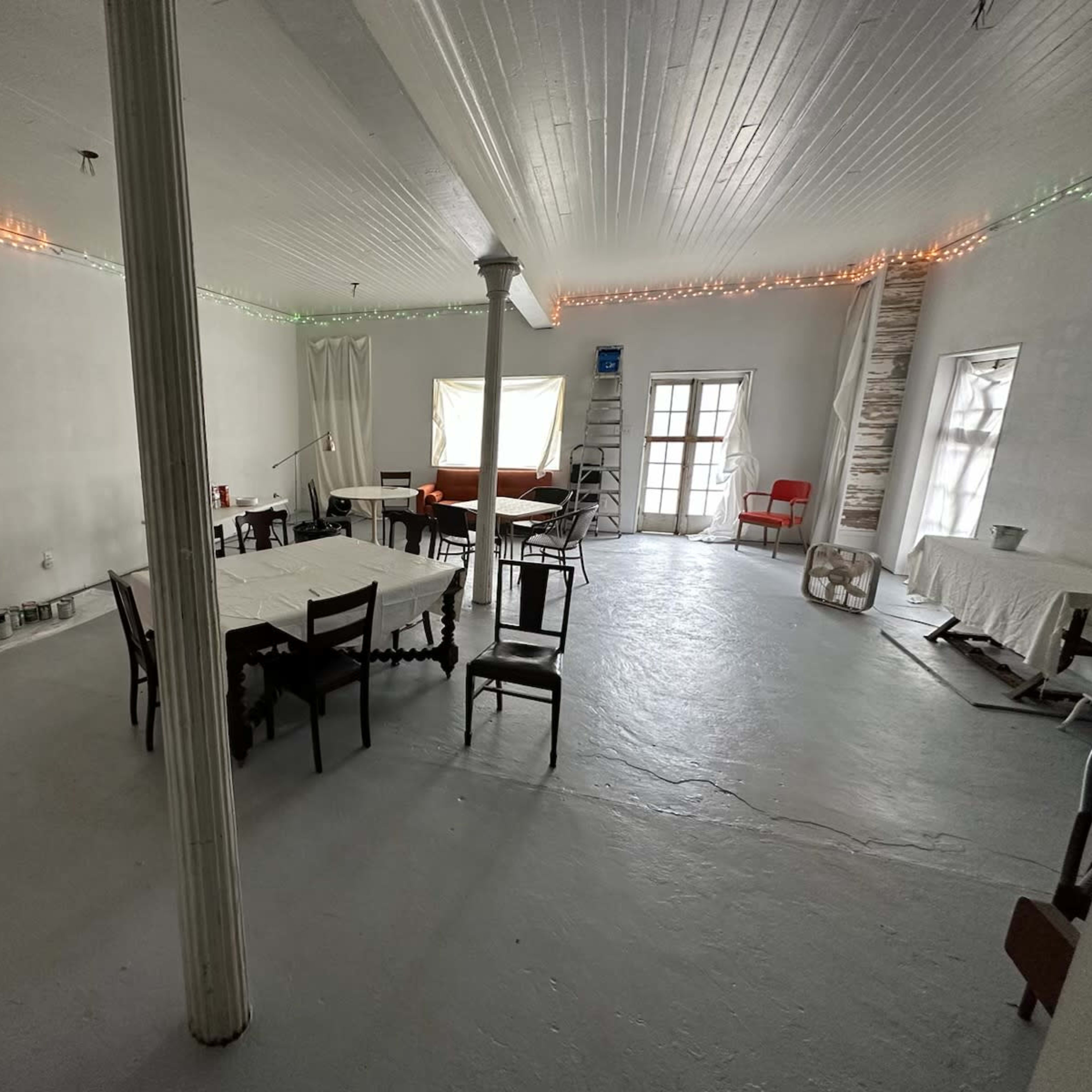 A spacious, uncluttered white room with several tables and chairs arranged, soft lighting along the walls, and large windows.