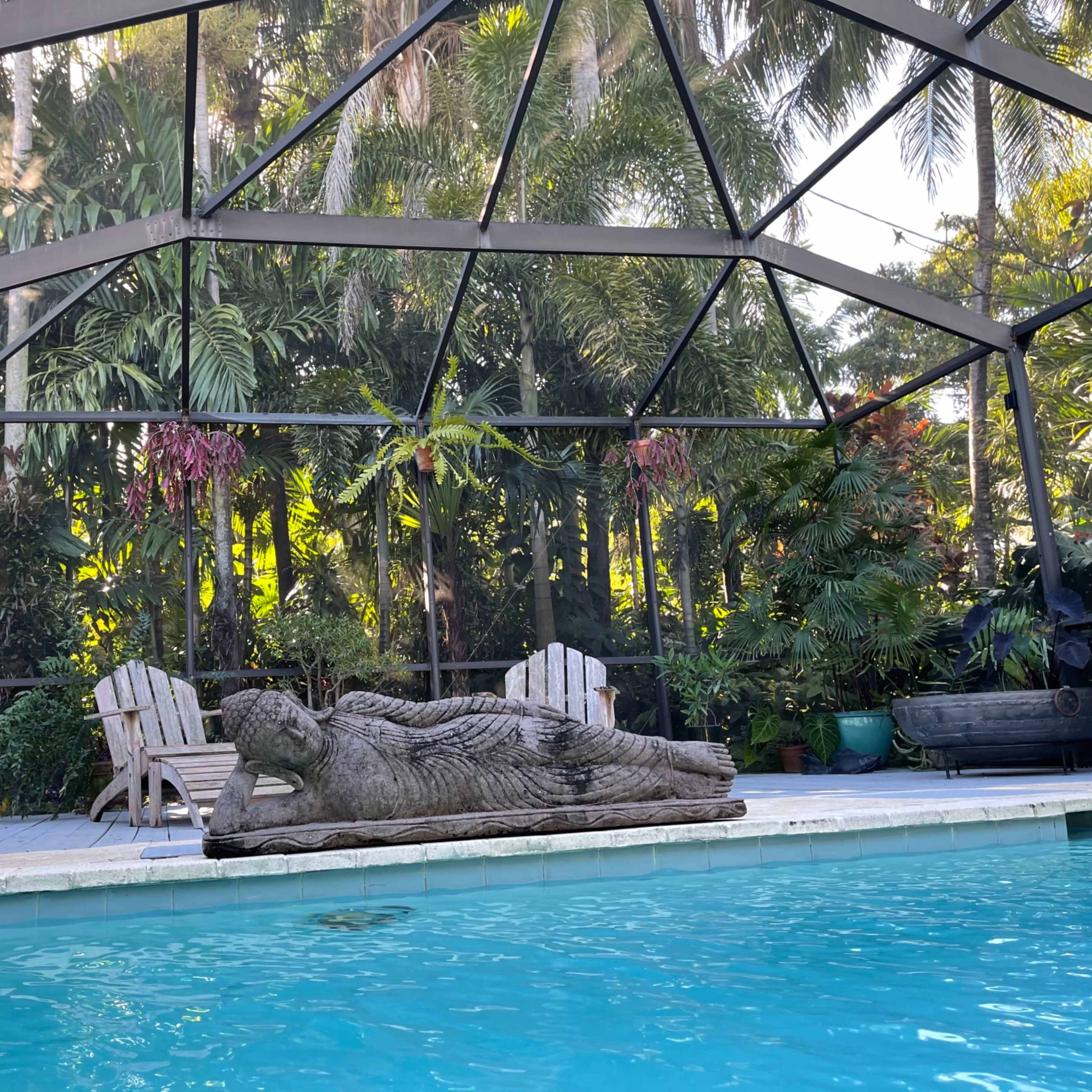 A large stone statue of a reclining figure rests beside a swimming pool, surrounded by lush tropical greenery and patio furniture.