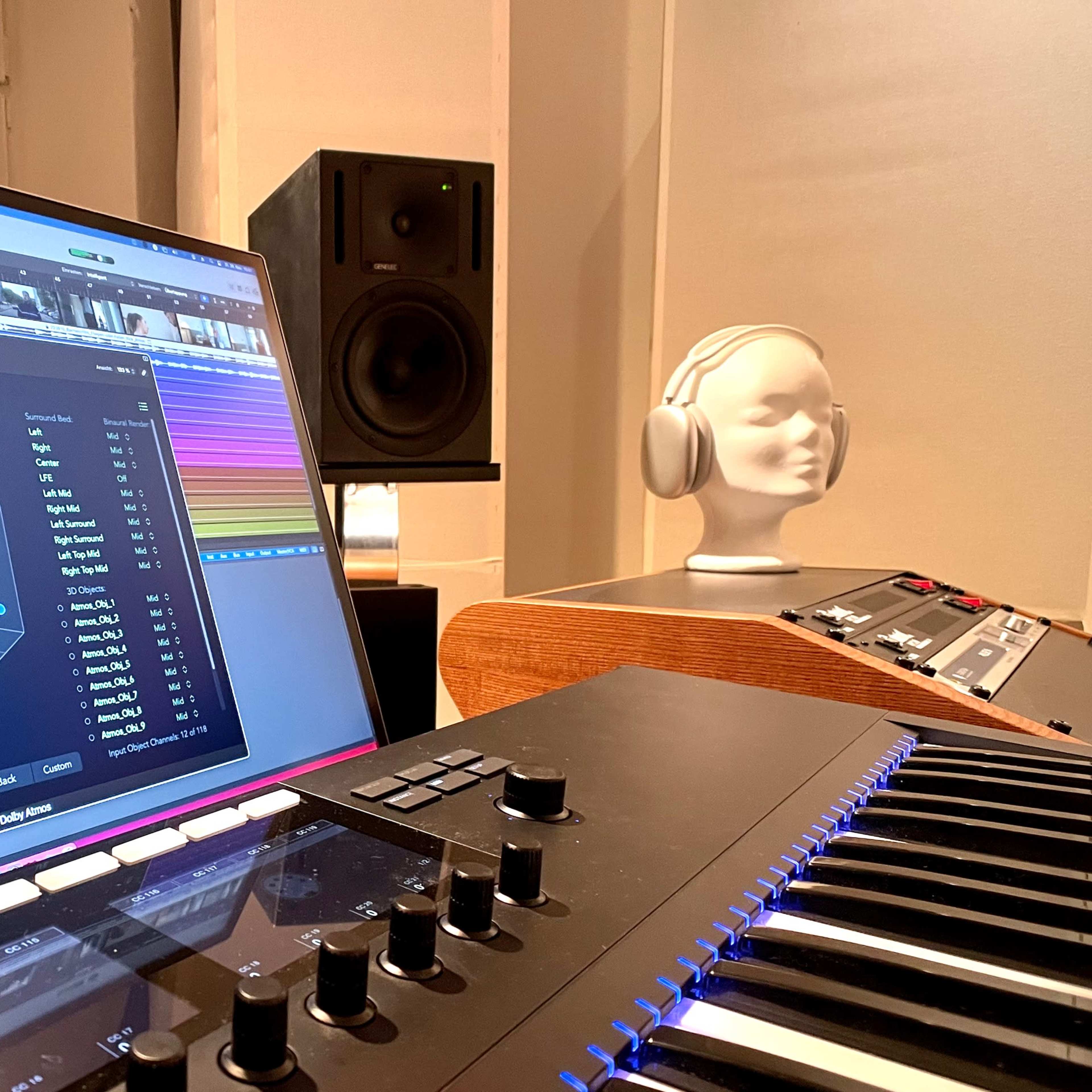A music production workspace featuring a computer screen displaying audio monitoring software, a MIDI keyboard, and a mannequin head with headphones positioned nearby.
