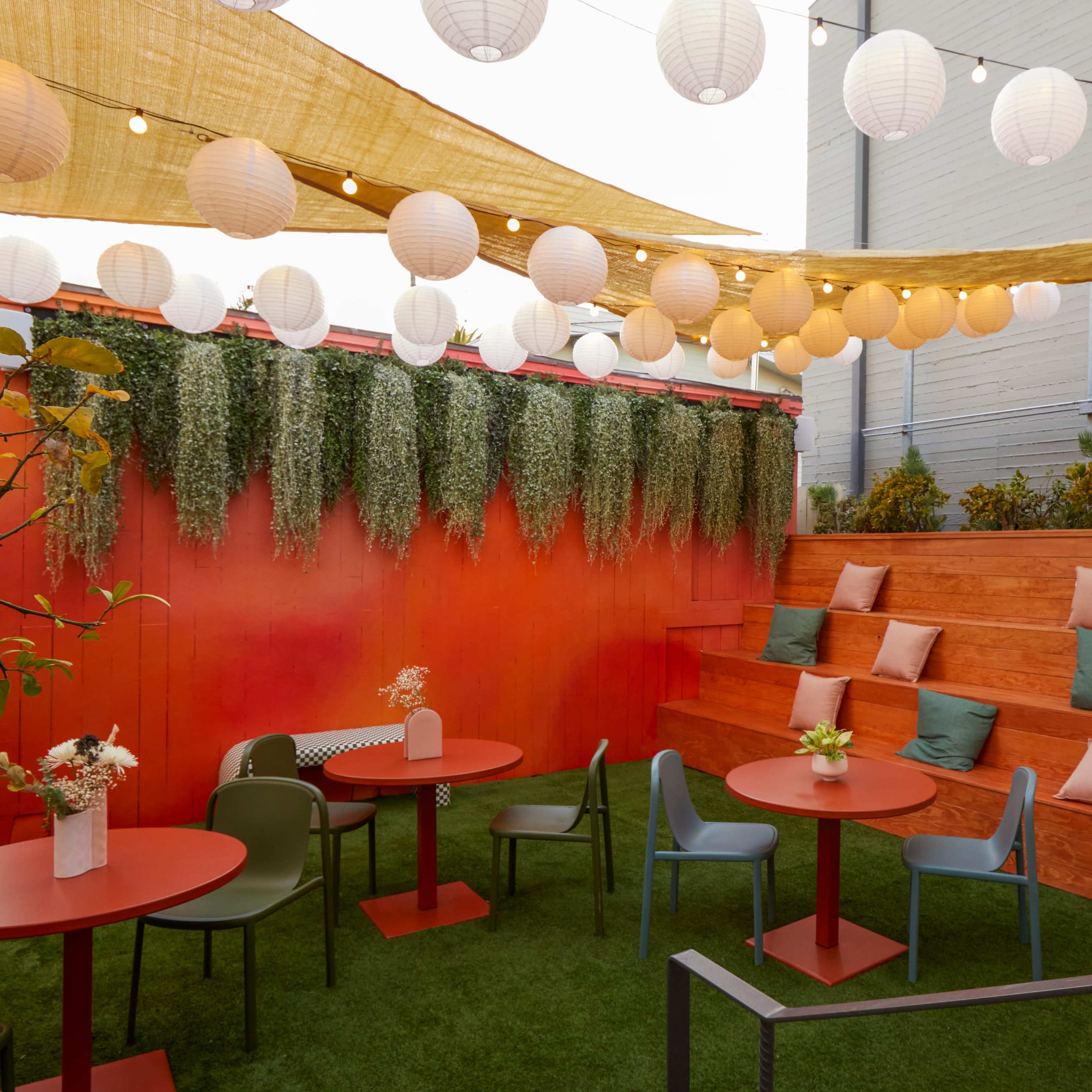 A vibrant outdoor seating area features wooden steps, round tables, and lanterns overhead, surrounded by greenery and decorative pillows.