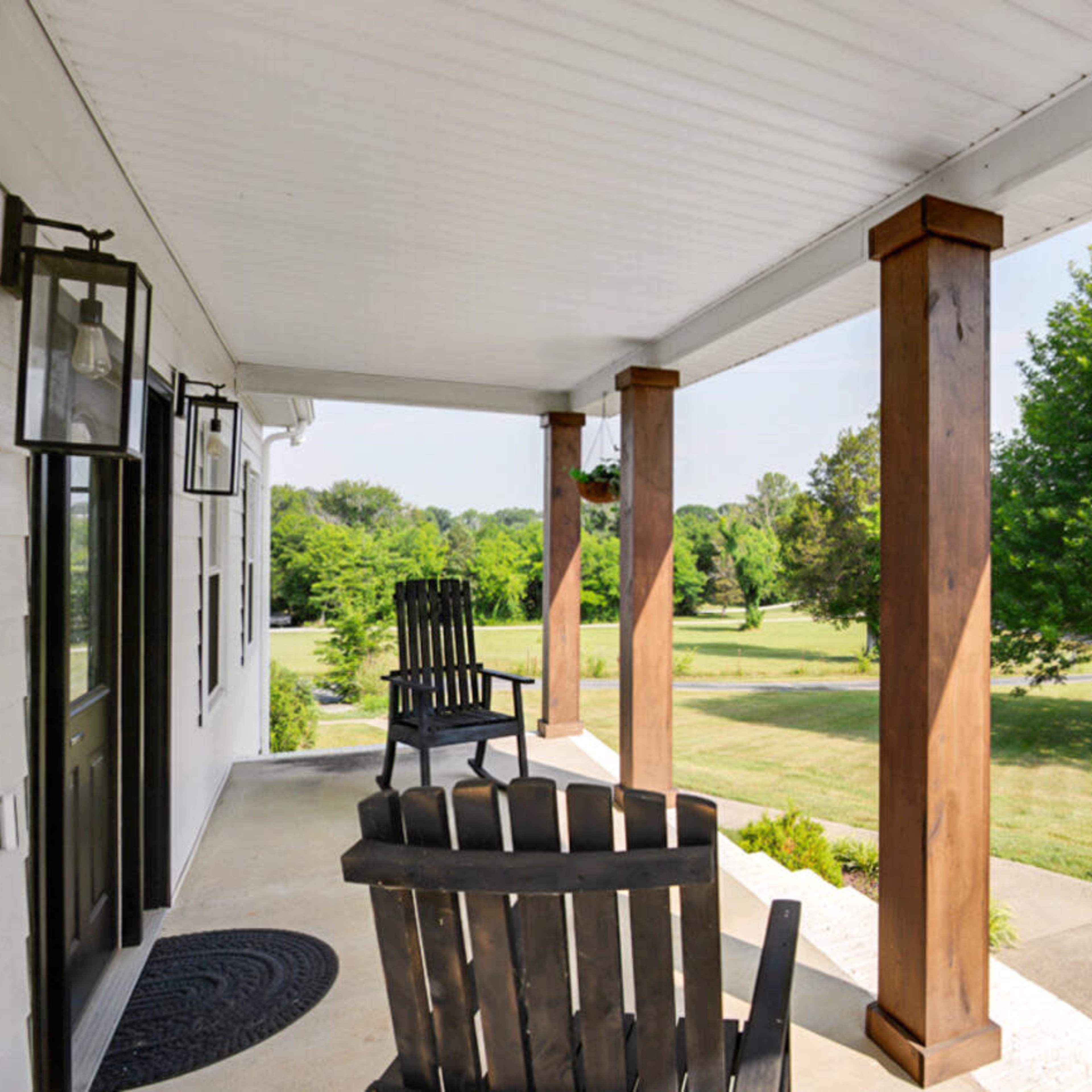 A front porch with two rocking chairs, wooden columns, and hanging planters, overlooking a green lawn and trees.