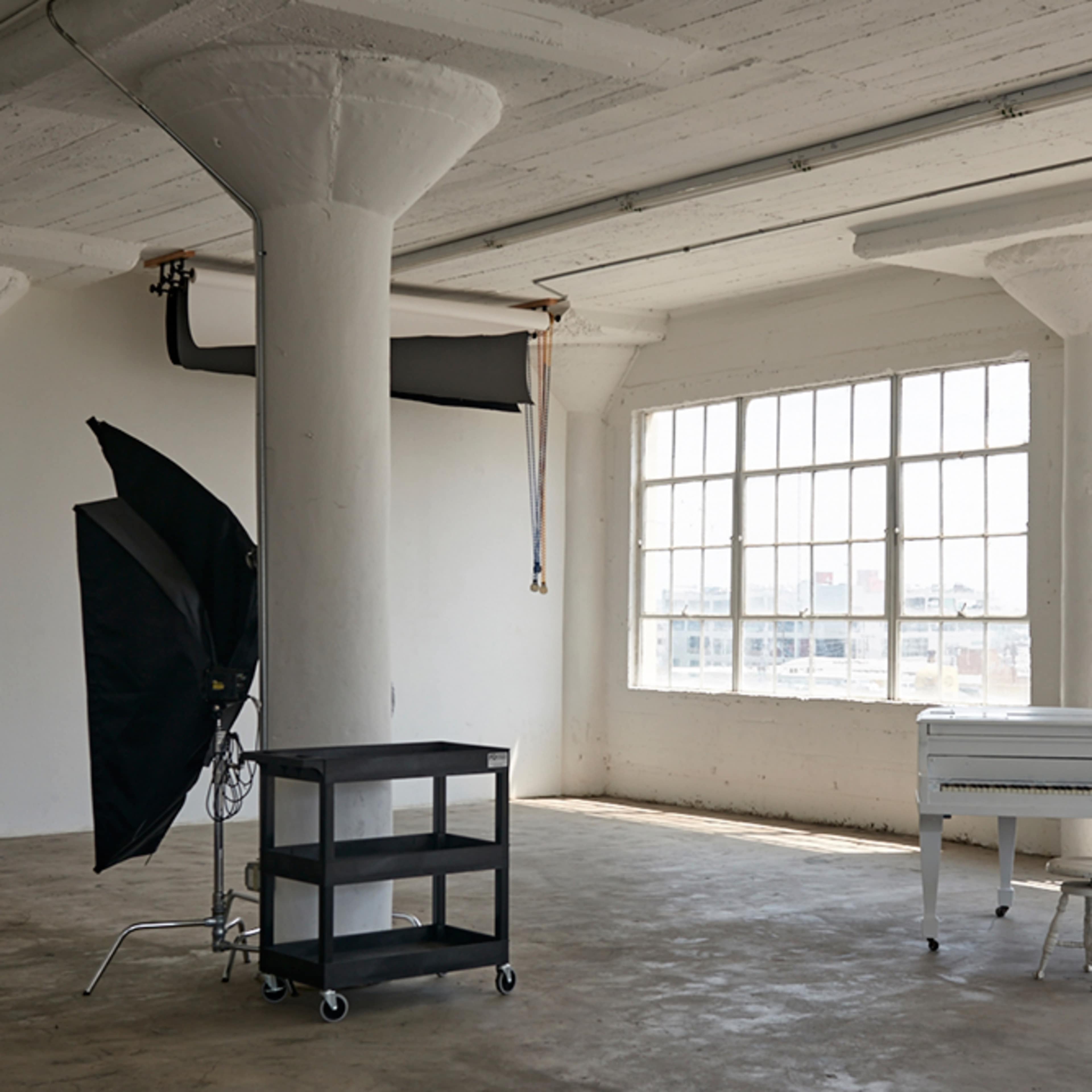A spacious, minimalist studio with large windows, a white piano, and a black photography umbrella on a cart.