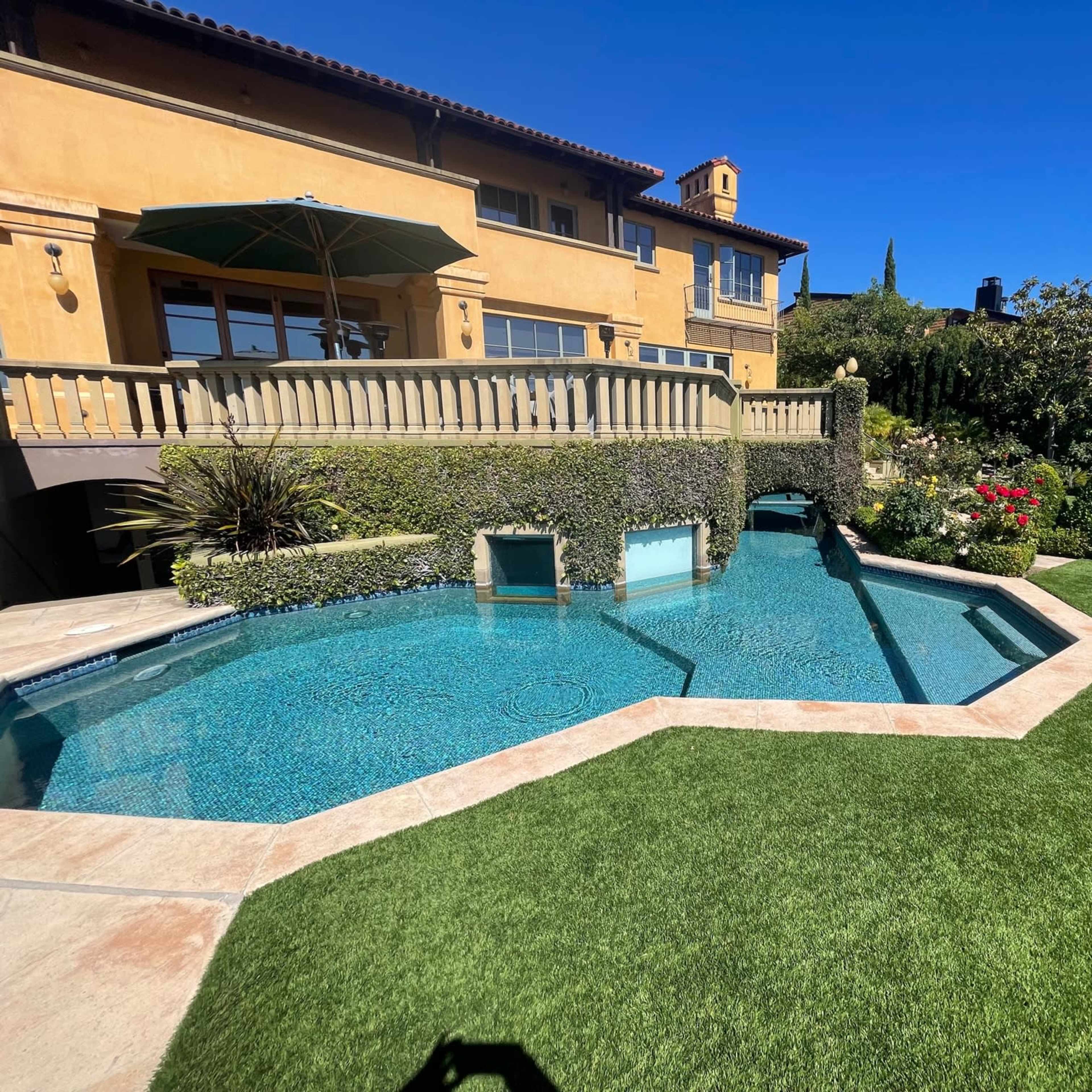 A landscaped backyard with a modern swimming pool, trimmed hedges, and a yellow stucco house under a clear blue sky.