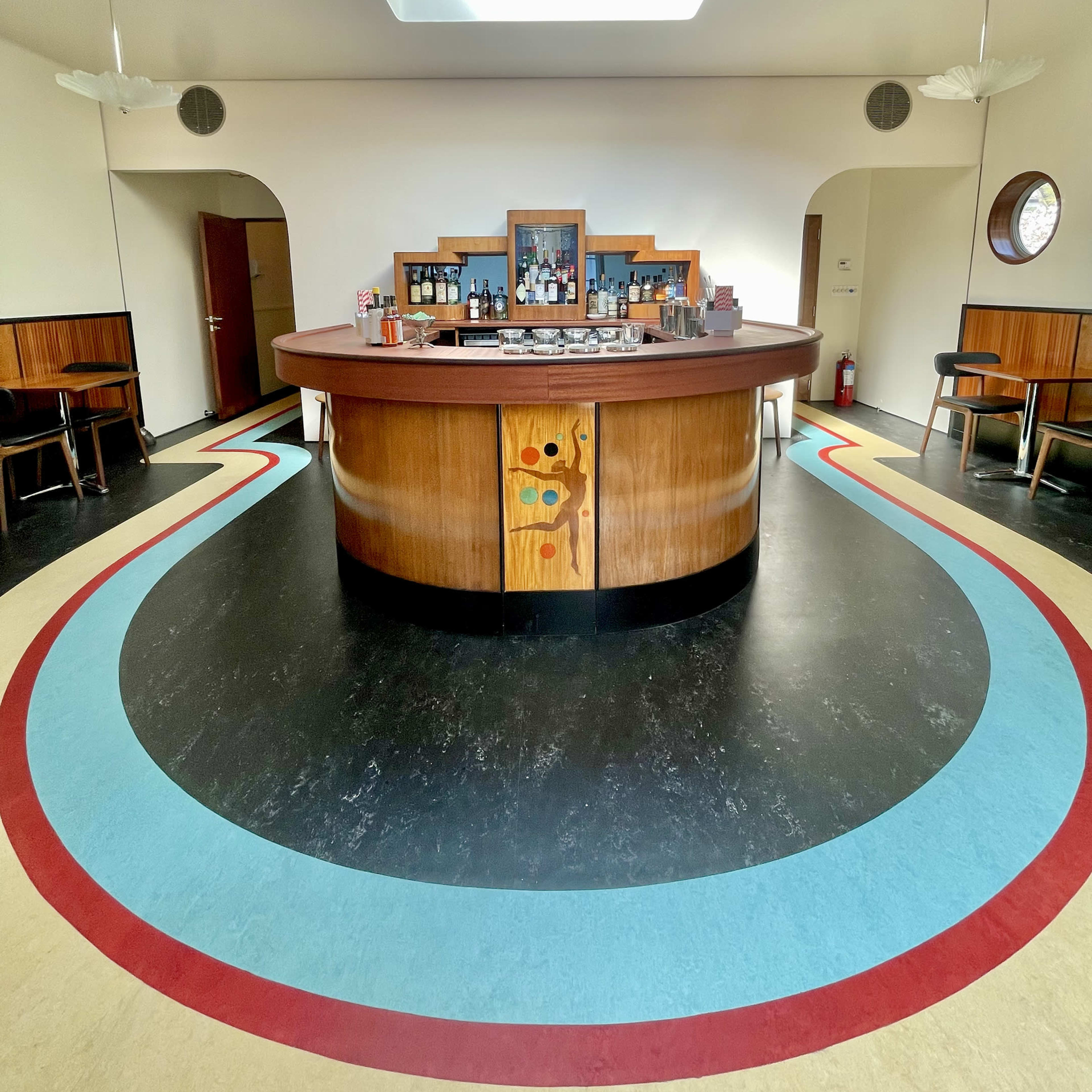 A circular wooden bar in a spacious interior with colorful curved flooring and a skylight above.