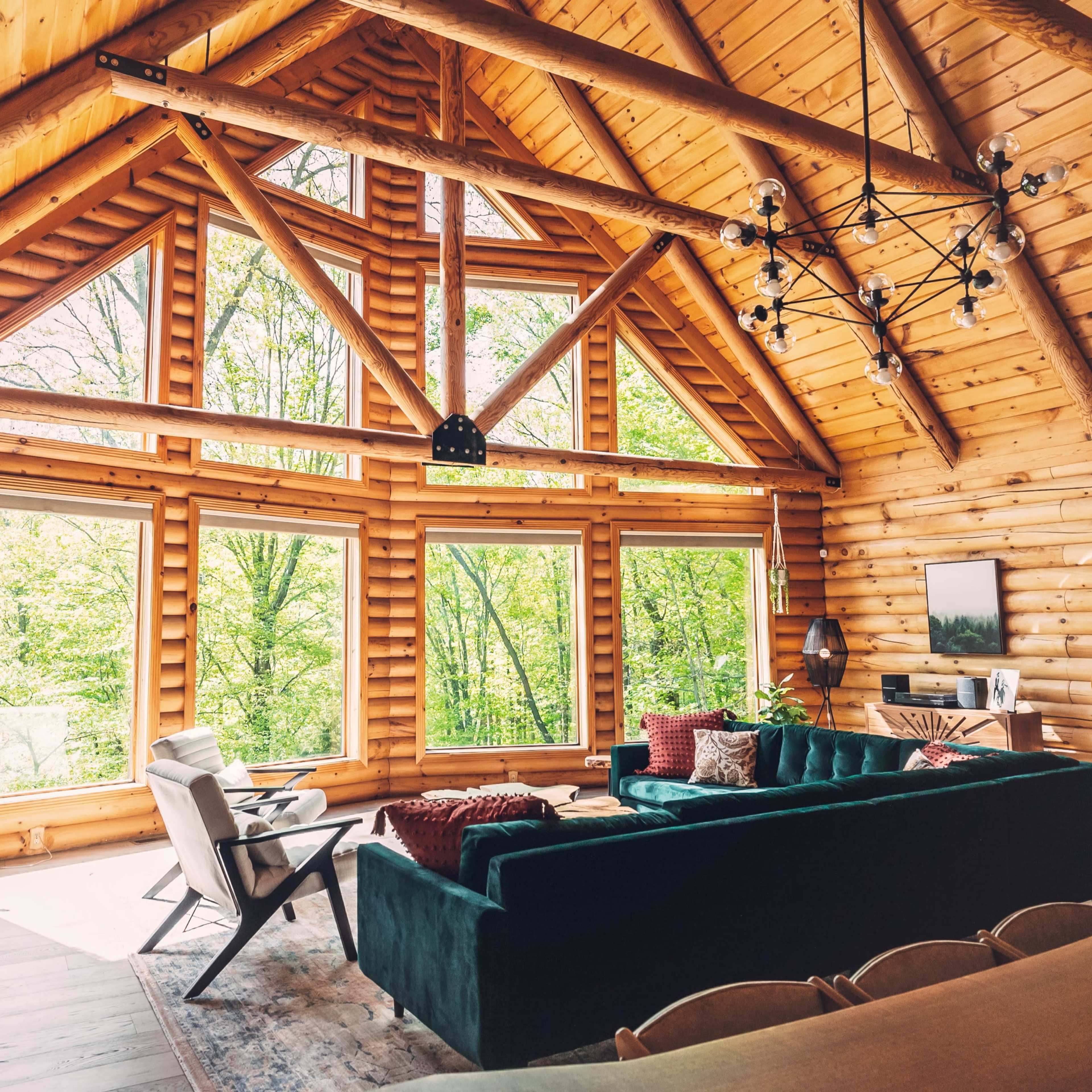 A spacious log cabin interior features large windows, a green sofa, and wooden beams, offering views of a forest.