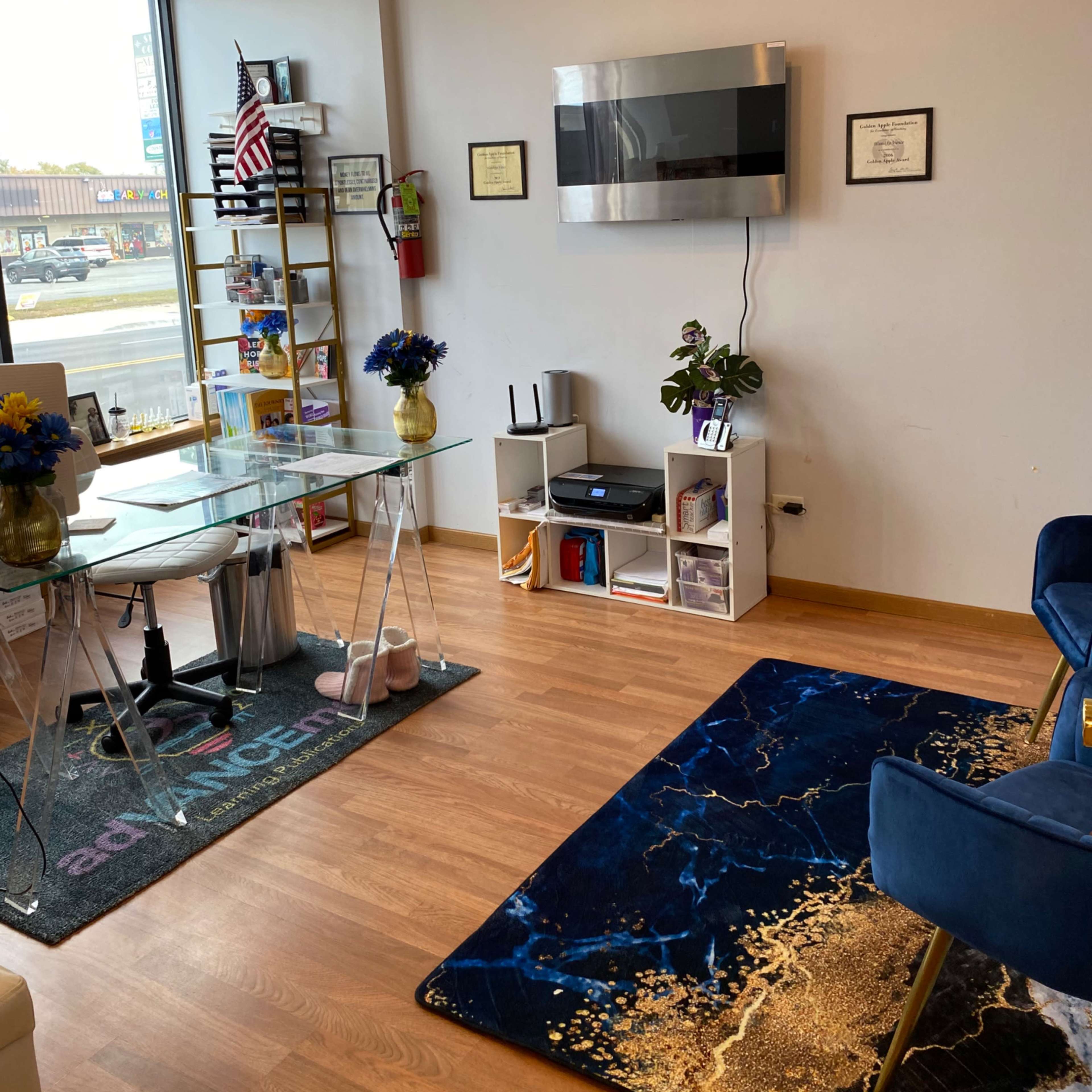 An office space featuring a glass dining table with chairs, a wall-mounted television, certificates framed on the wall, and a blue rug on the wooden floor.
