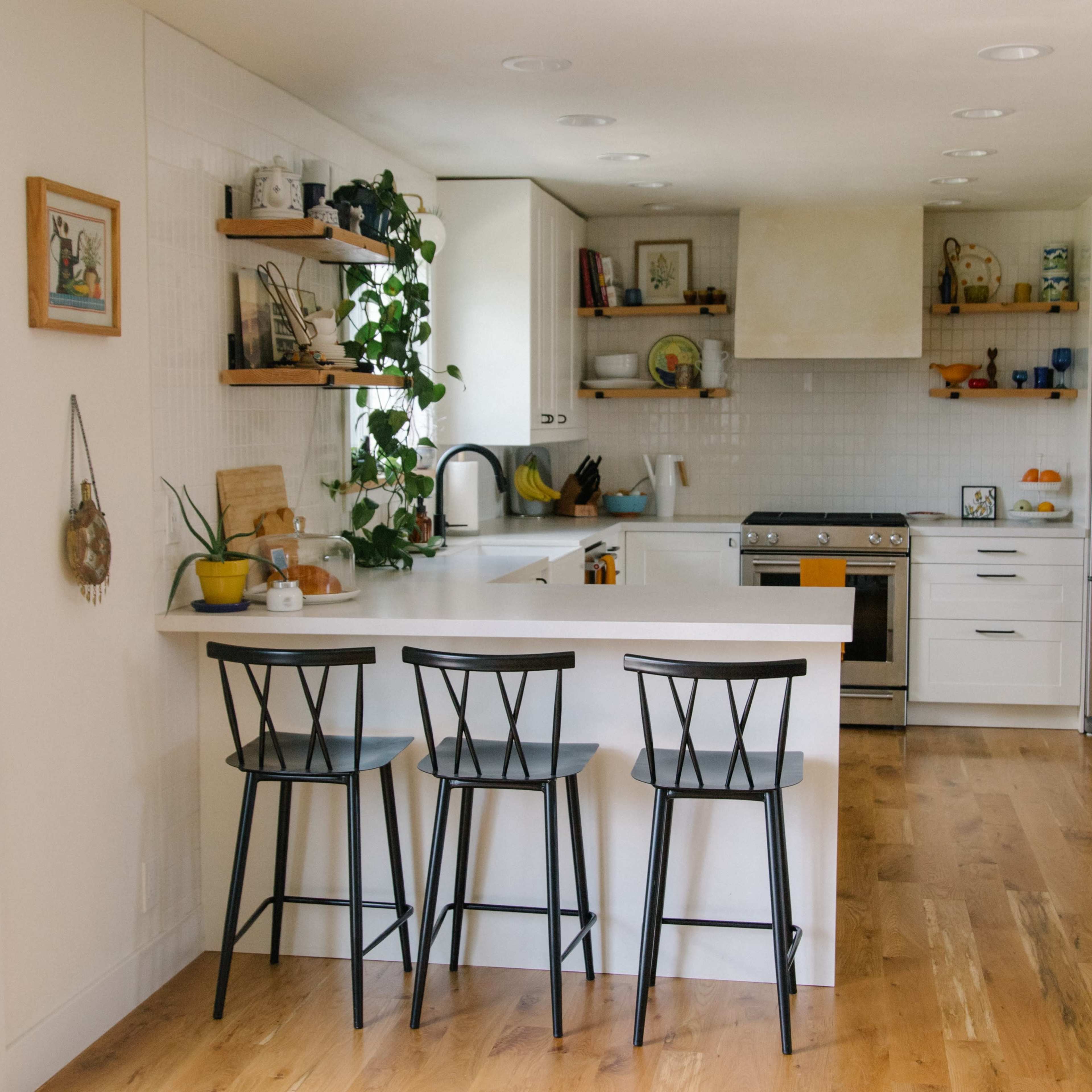 A modern kitchen features a central island with three black bar stools, white cabinetry, and natural wood flooring.