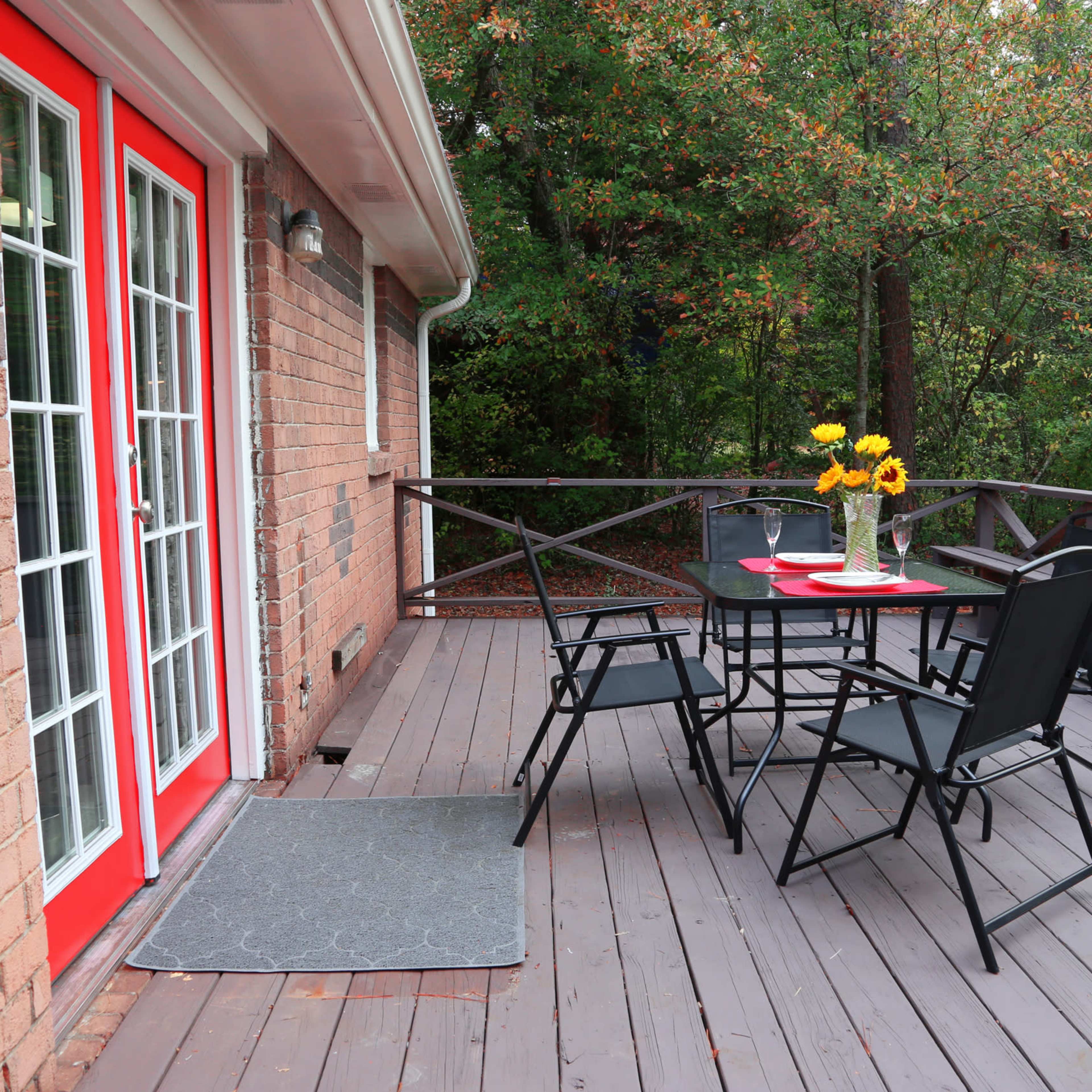 A wooden deck with a table and four chairs, featuring a vase of sunflowers, alongside a brick house with red French doors.