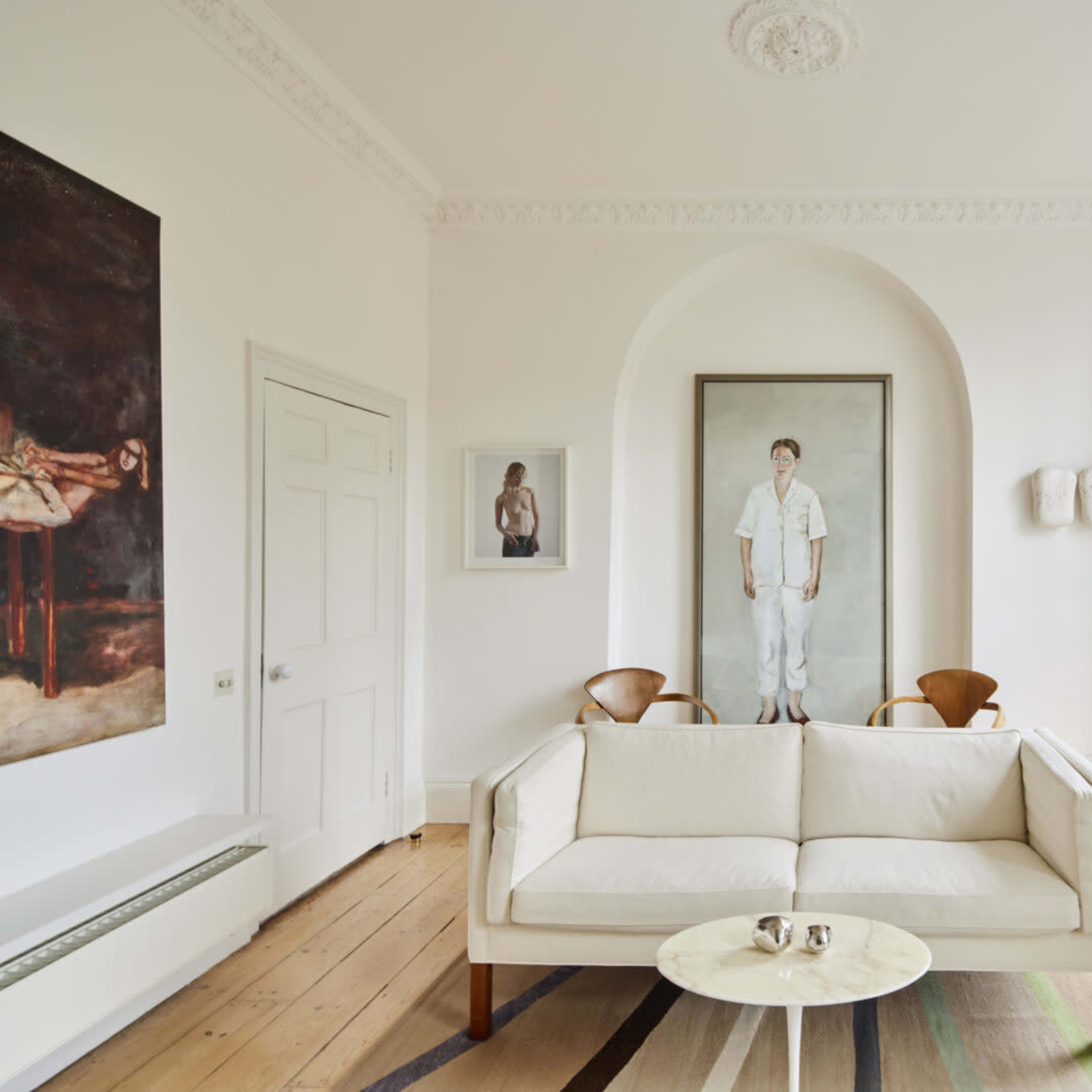 A bright living room featuring a white sofa, two wooden chairs, and framed artworks on the walls.