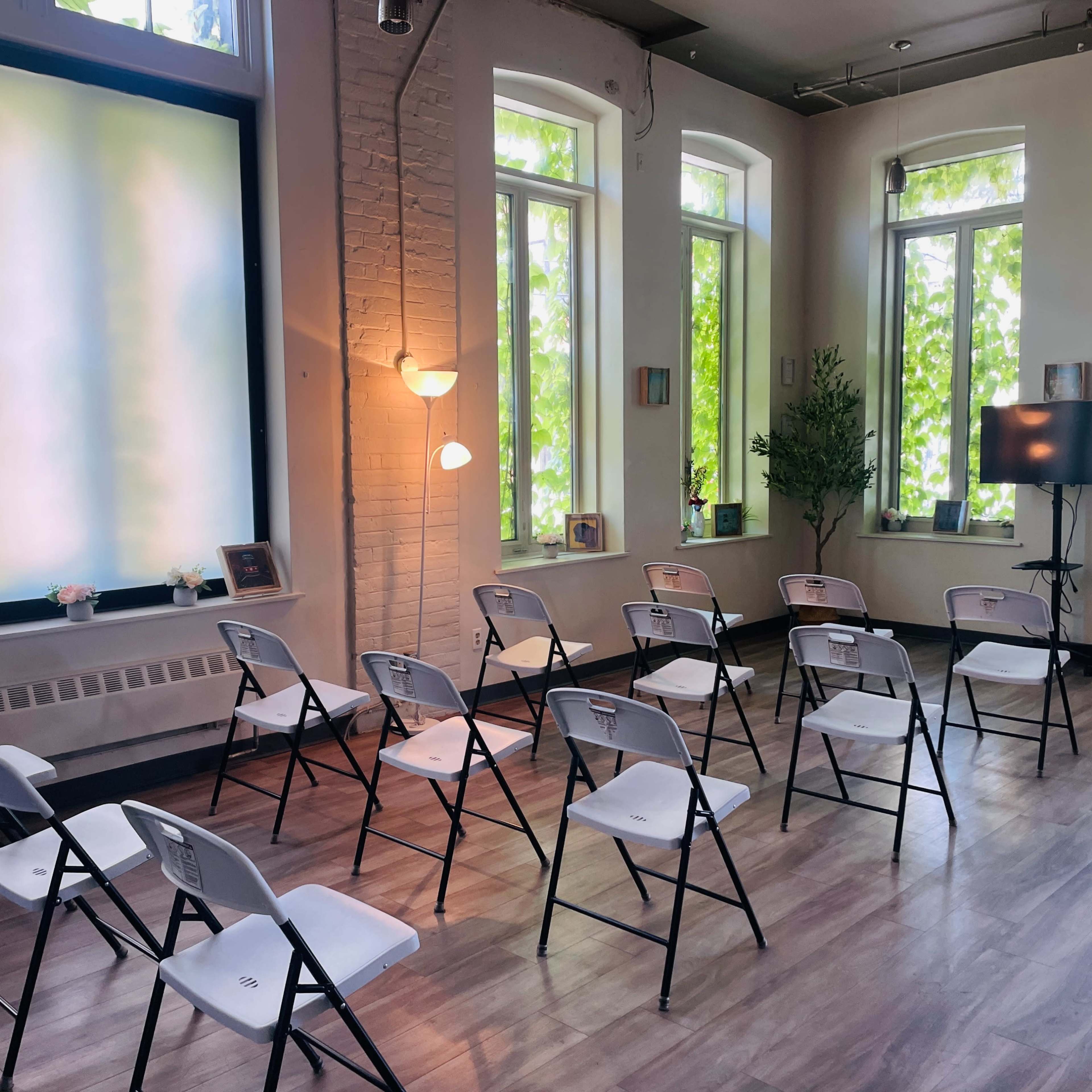 A small, bright room arranged with white folding chairs facing a TV, surrounded by large windows with greenery outside.