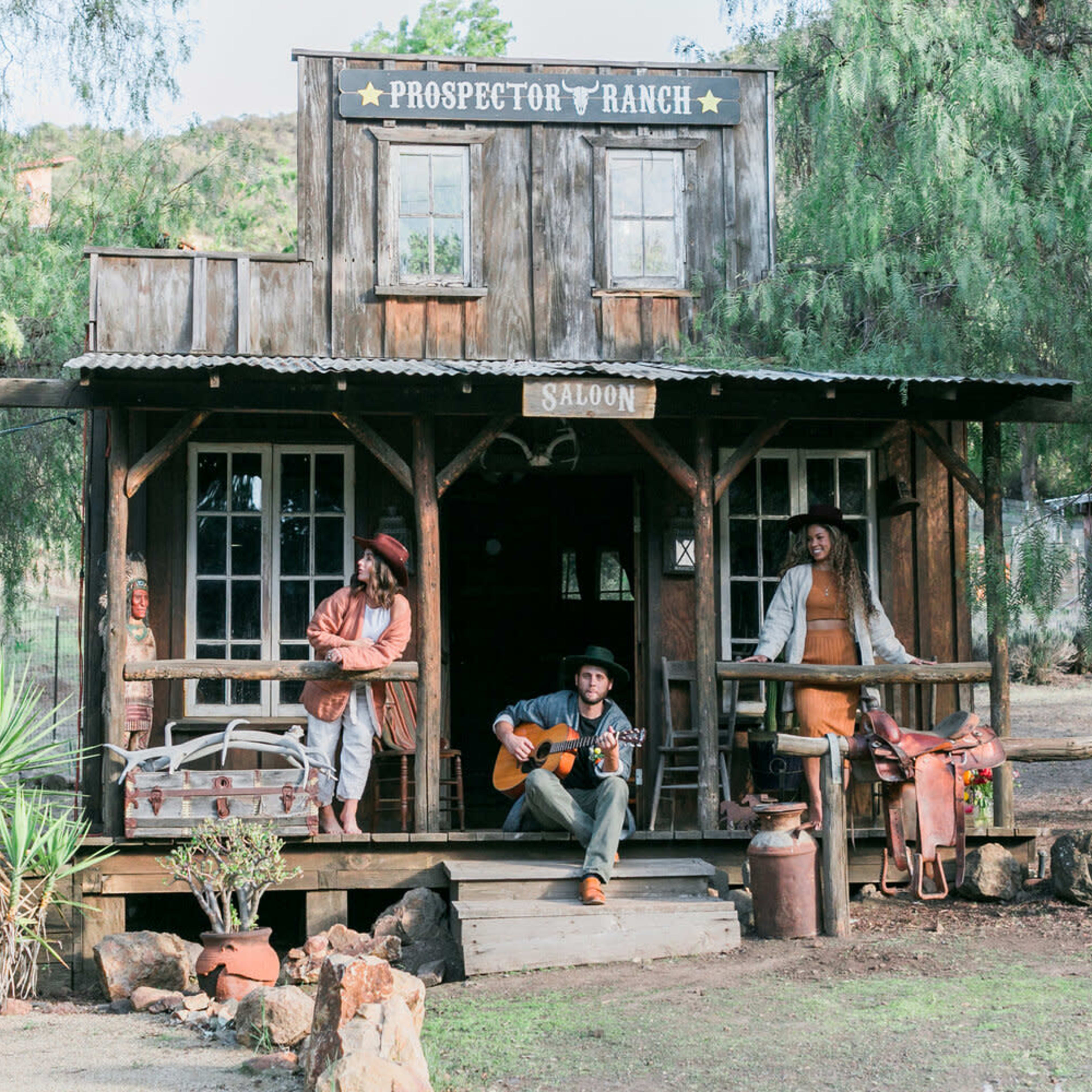 A cowboy sits on the steps of a wooden saloon, playing guitar, while two women pose on the porch at Prospector Ranch.