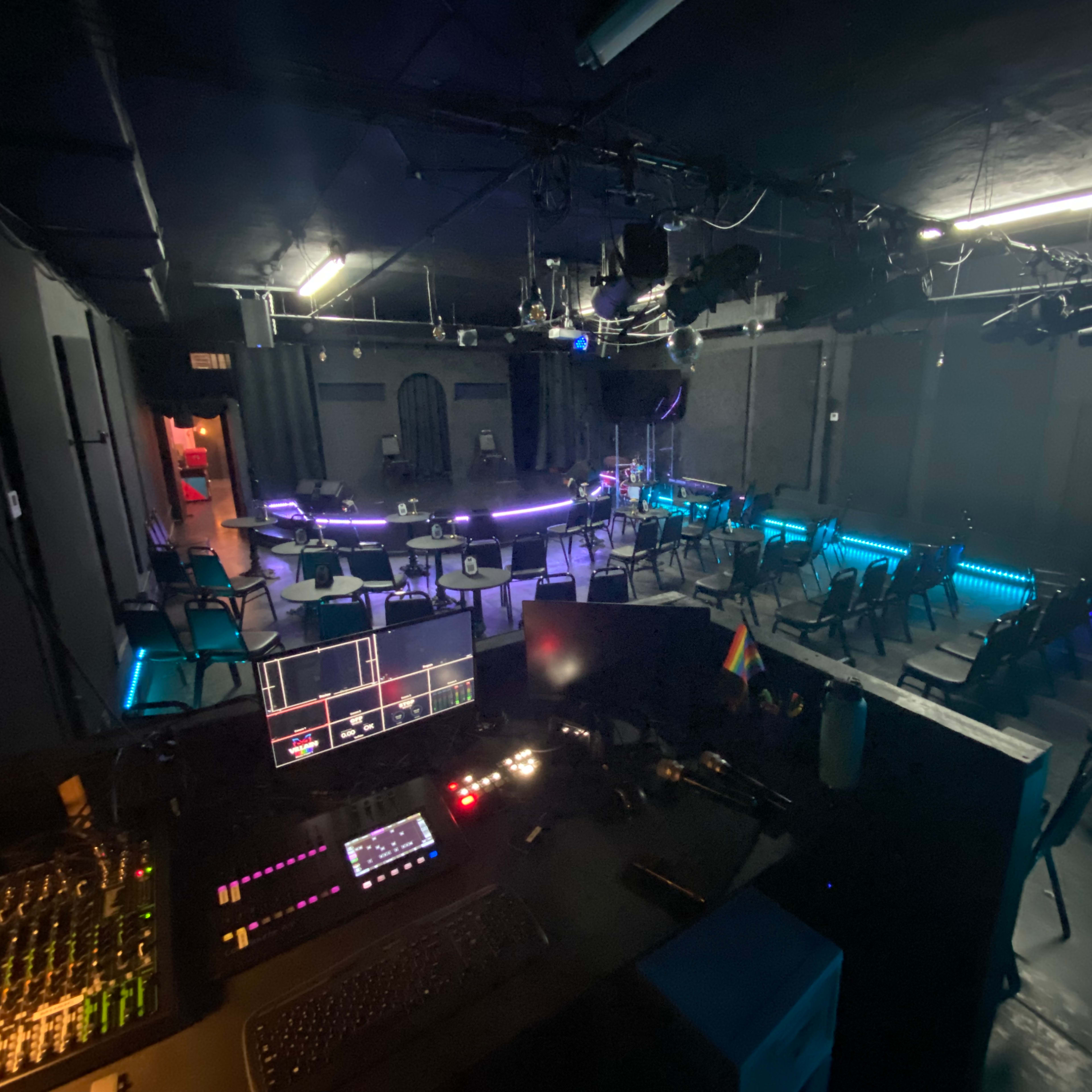 A dimly lit performance space with a soundboard in the foreground, featuring rows of chairs and a circular stage illuminated by blue lights.
