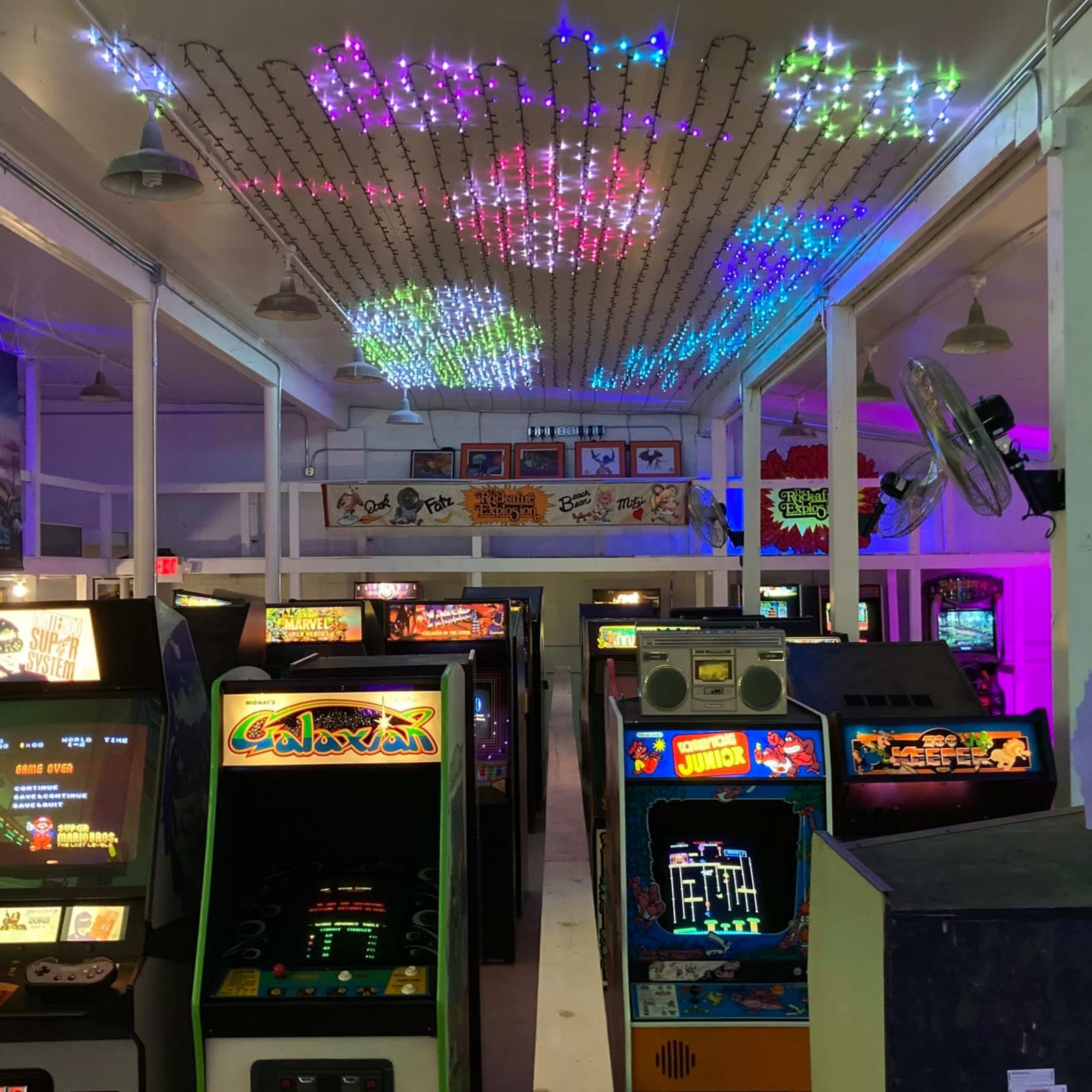 A row of vintage arcade games under a ceiling adorned with colorful lights in a nostalgic gaming venue.