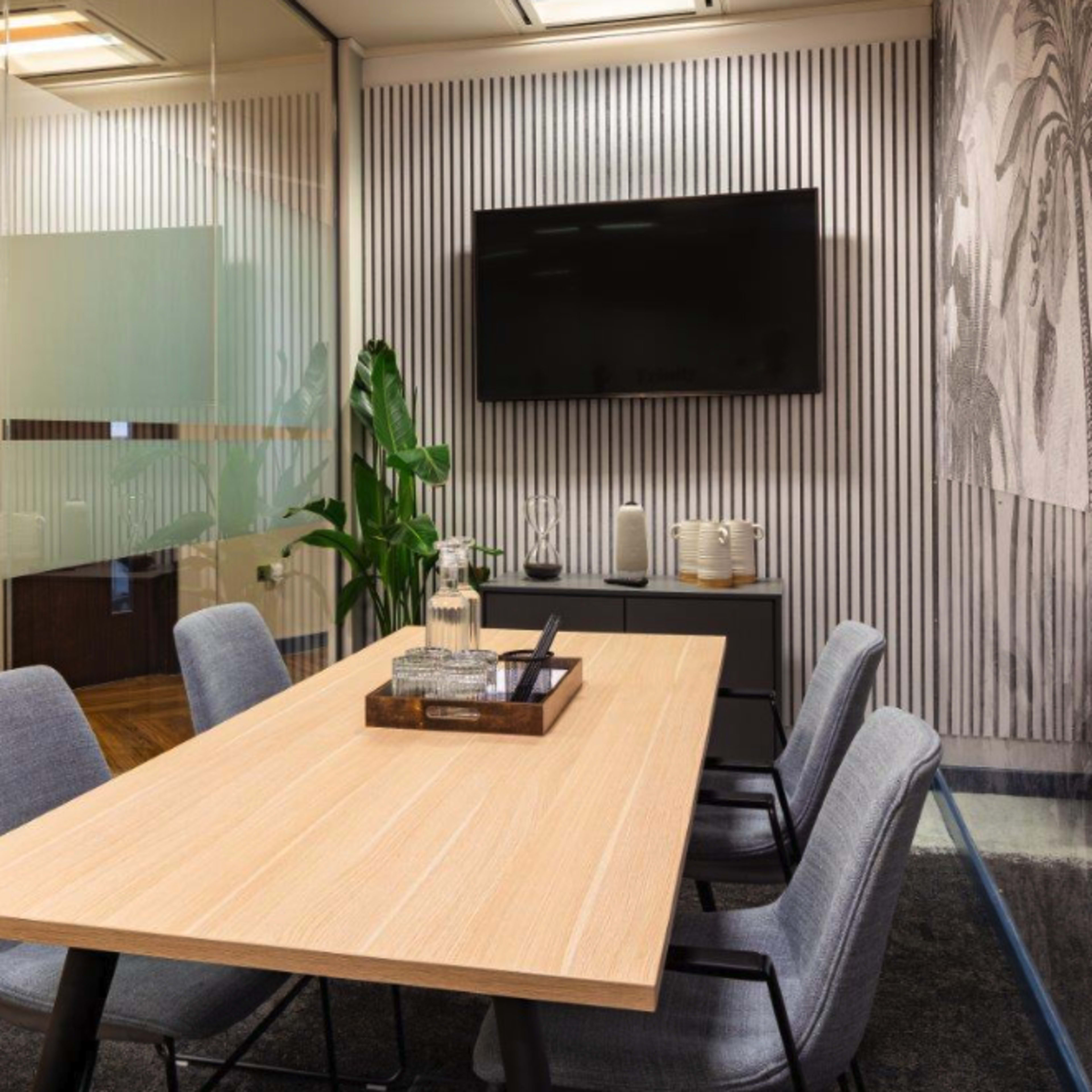 A modern conference room features a wooden table surrounded by gray chairs, with a television mounted on the wall and a decorative plant nearby.