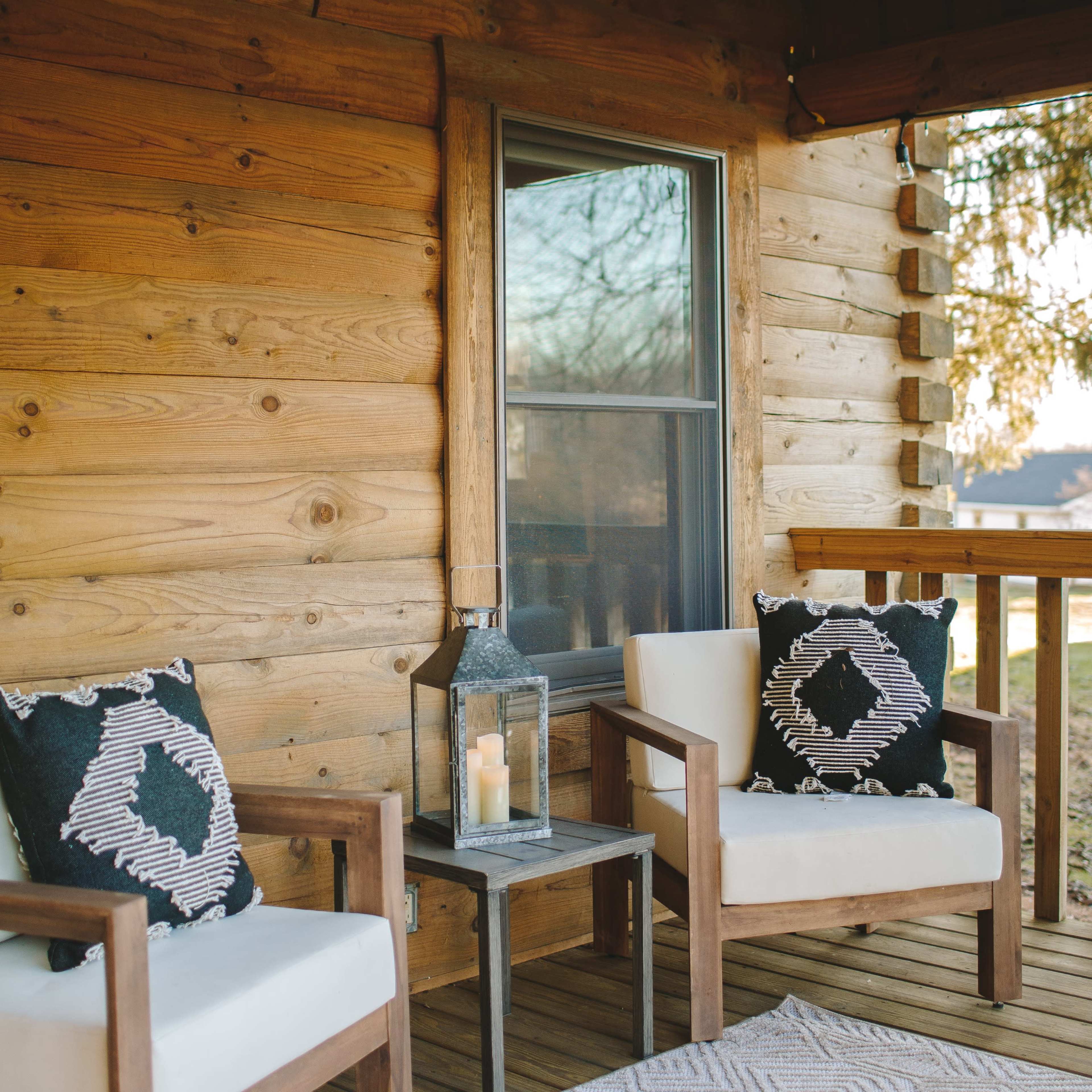 A rustic porch features two wooden chairs with decorative pillows, a small table holding a lantern and a candle, and a window in the wood-paneled wall.