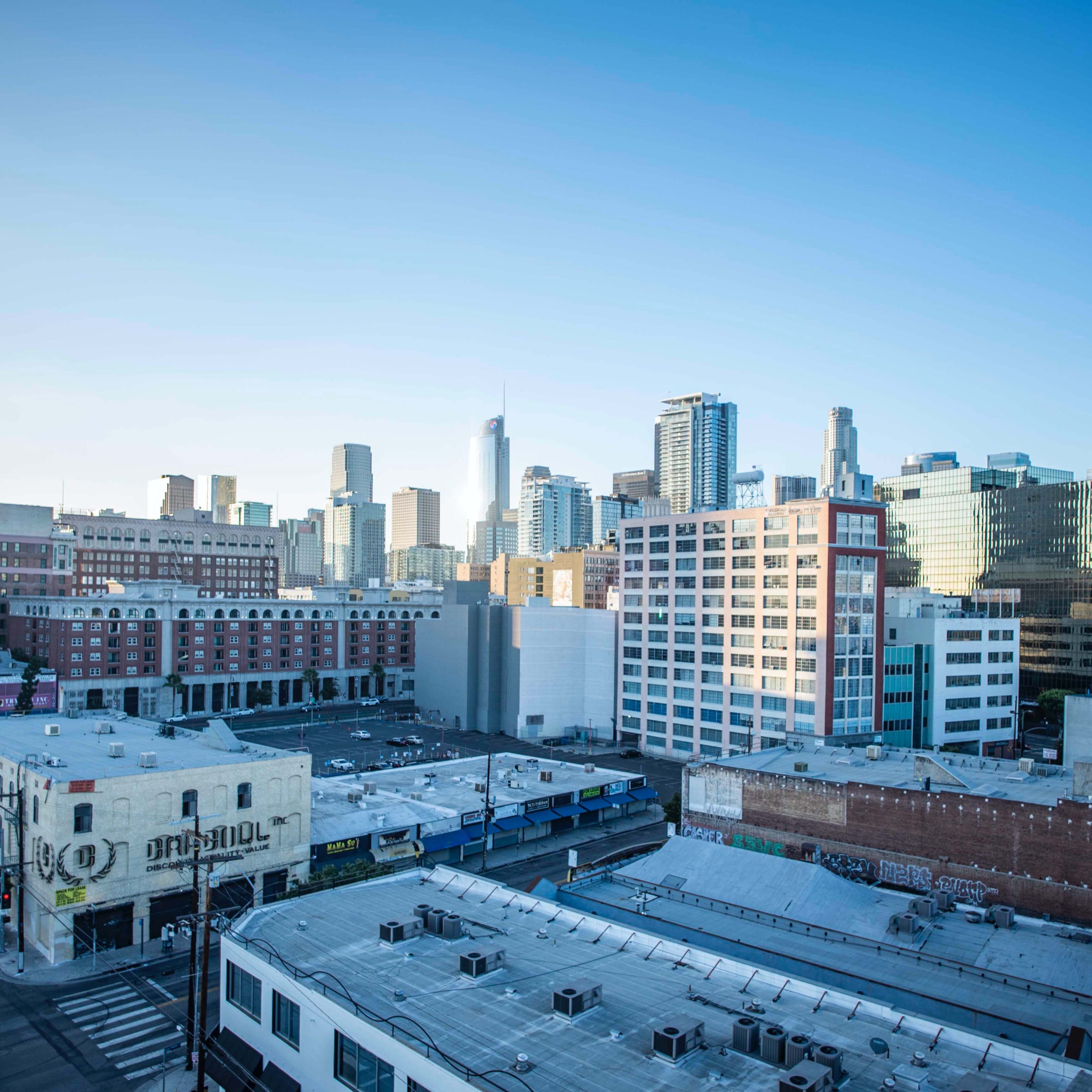 A skyline of tall buildings in a city, with a clear blue sky above and a street lined with low-rise structures in the foreground.