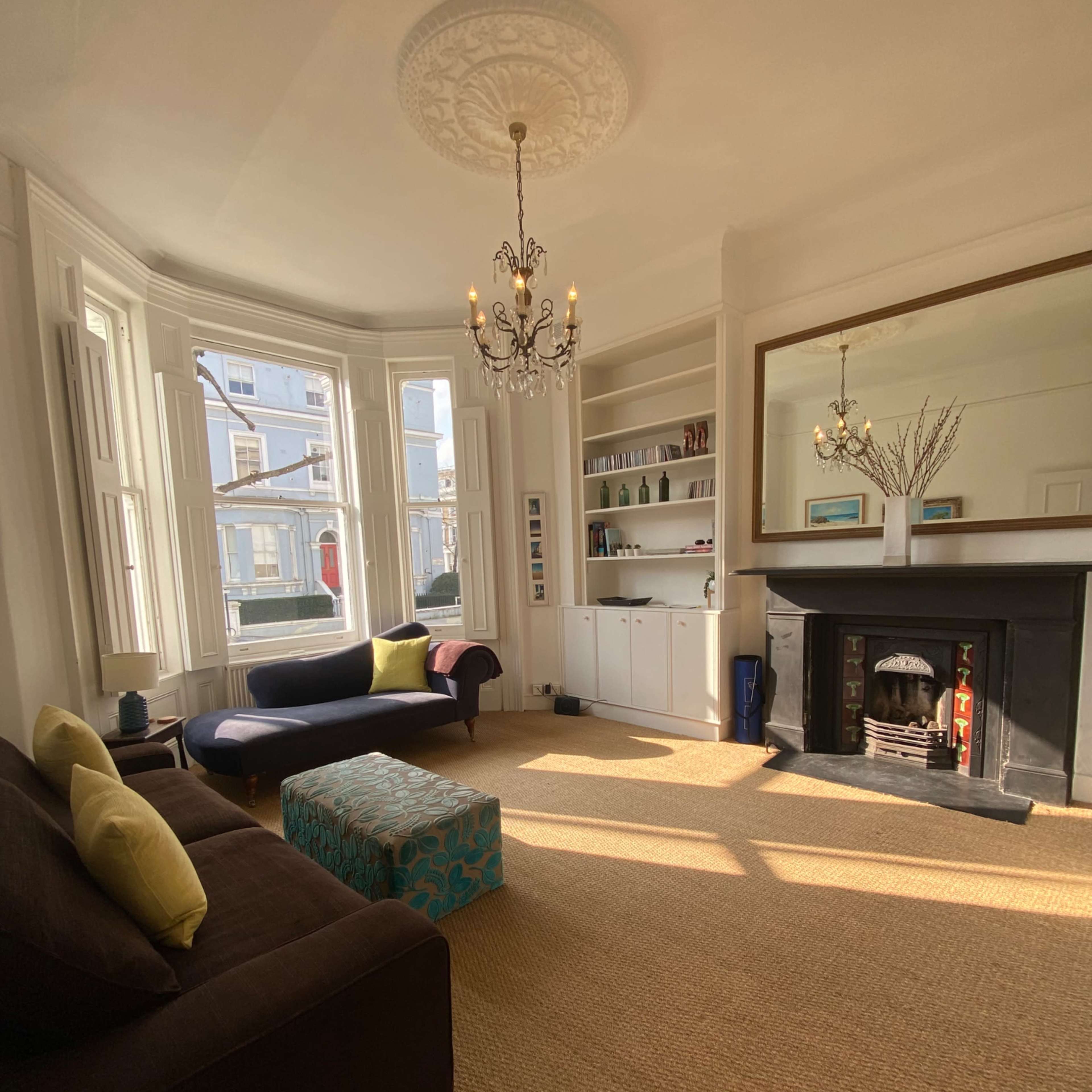 The living room features a bay window, a fireplace, a brown sofa with bright cushions, and a blue patterned ottoman.