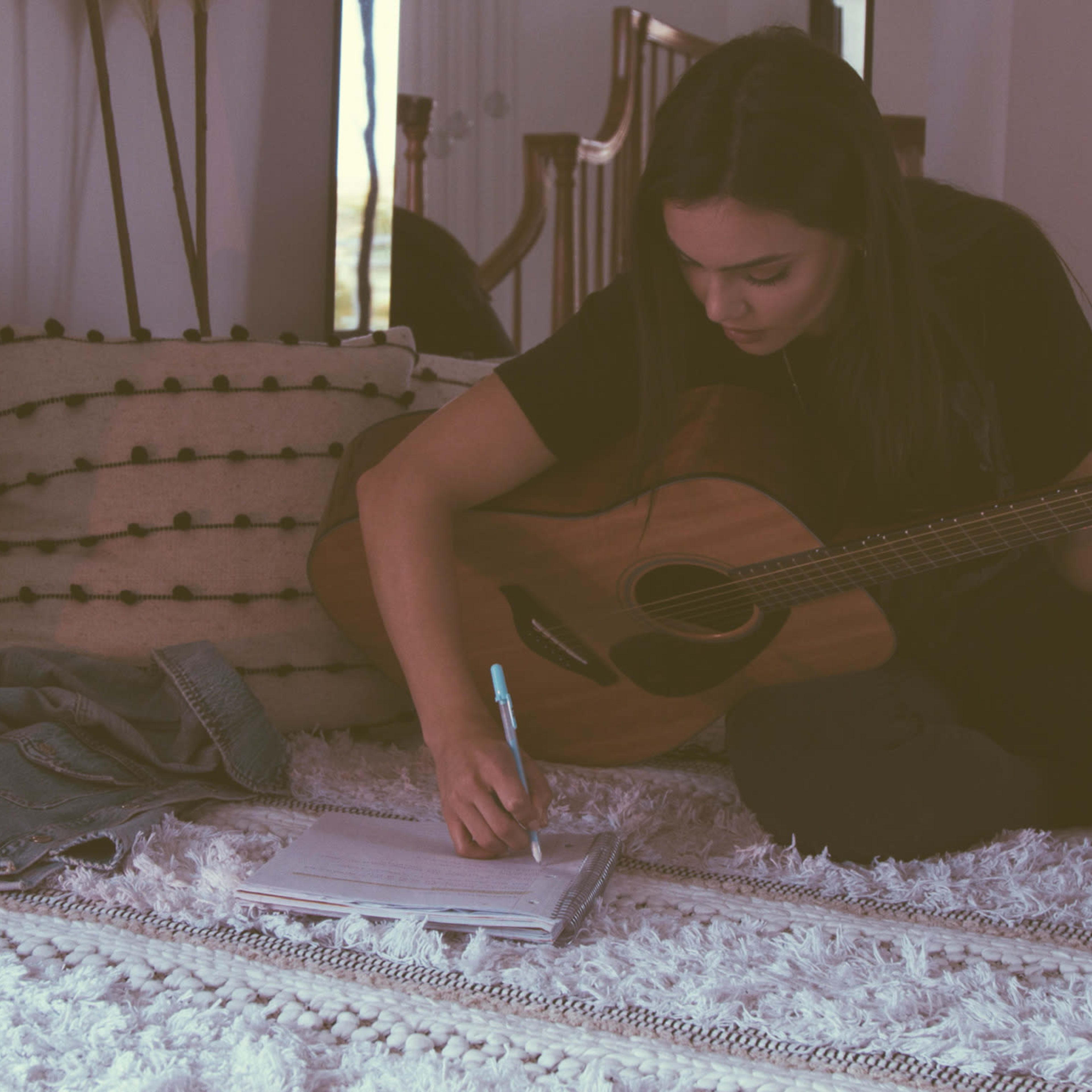 A person sits on a rug with a guitar in their lap while writing in a notebook.