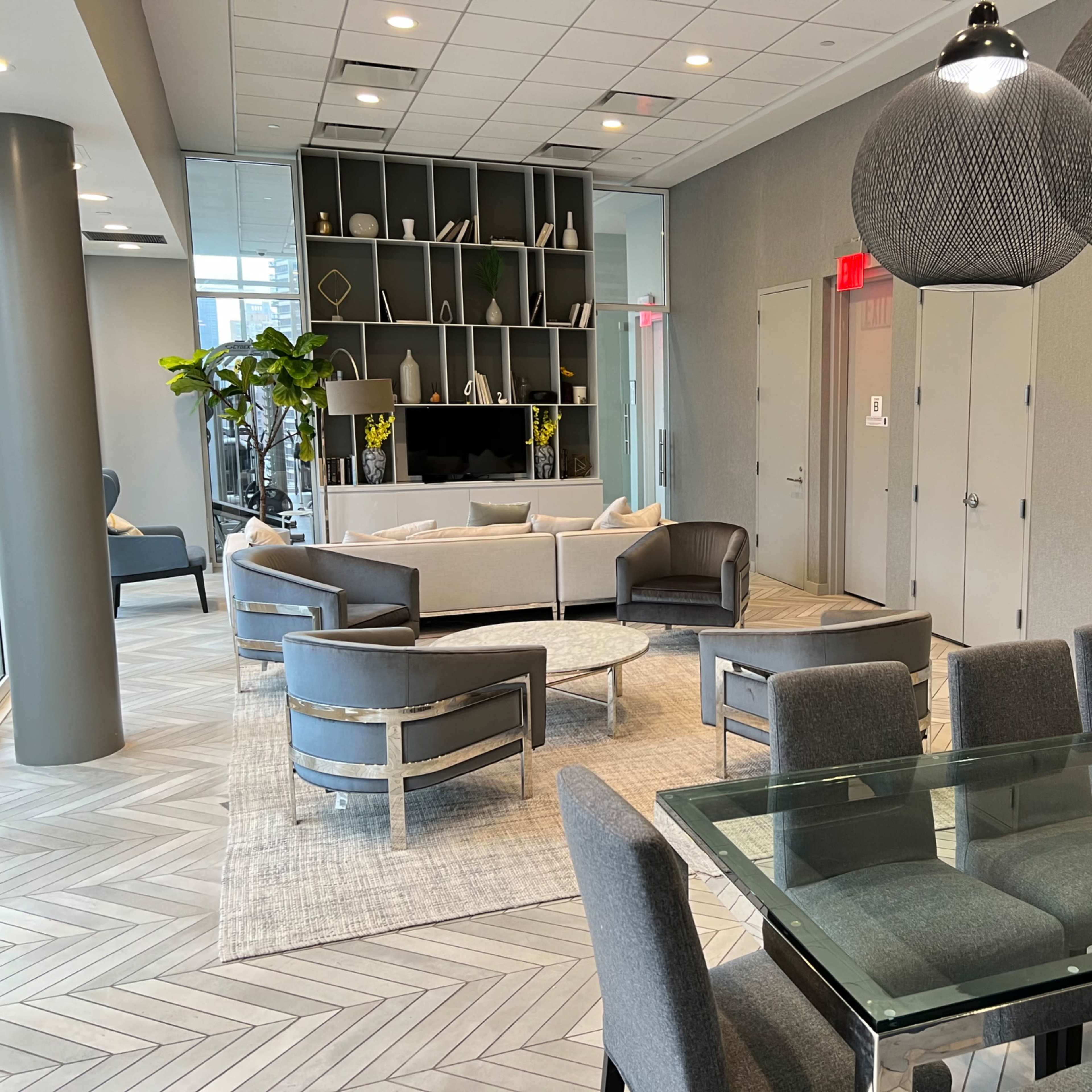 A modern lounge area with a seating arrangement of gray chairs and a sofa, a glass dining table, and shelves filled with decorative items.