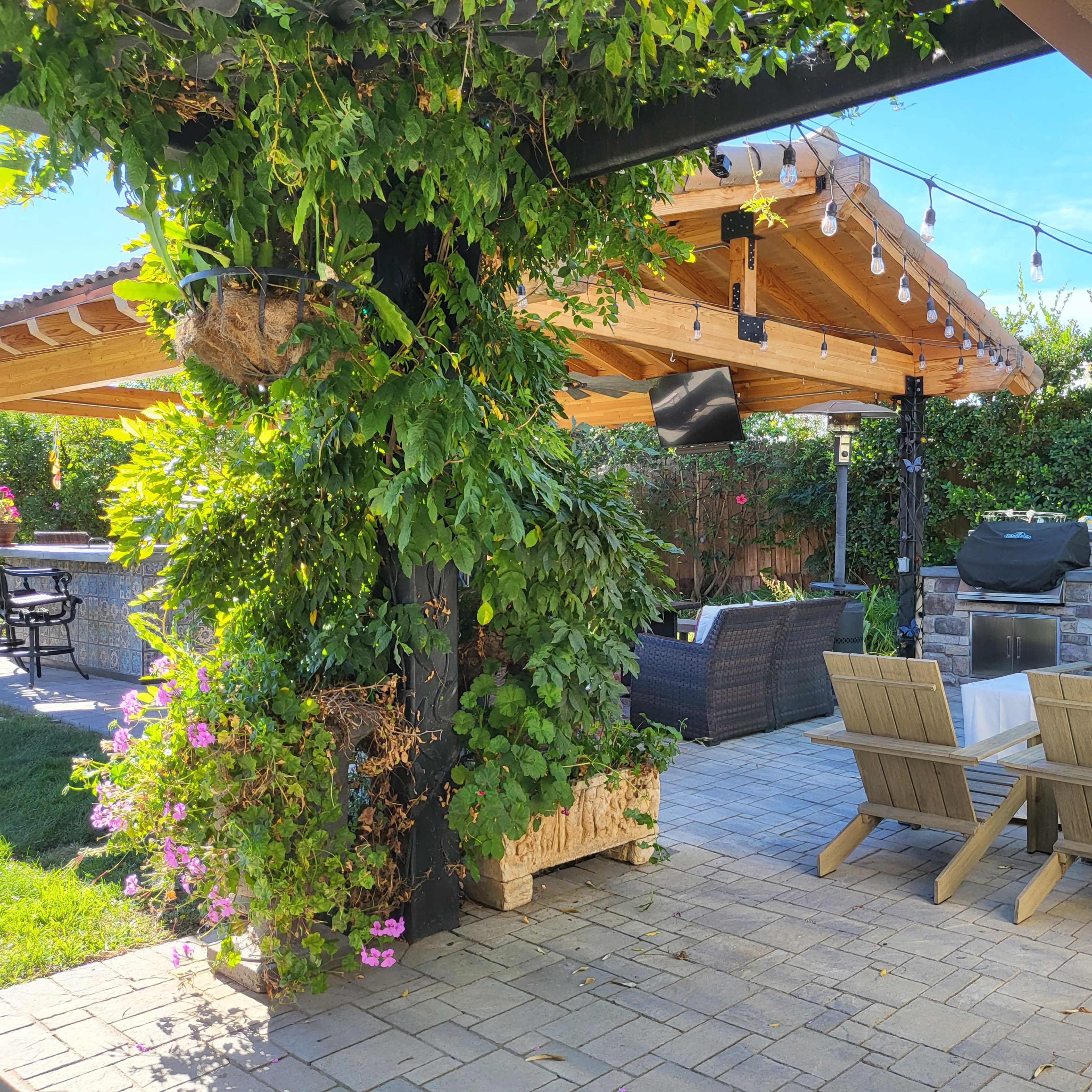 An outdoor patio area featuring a covered seating space with wooden furniture, a barbecue grill, and lush greenery surrounding the space.