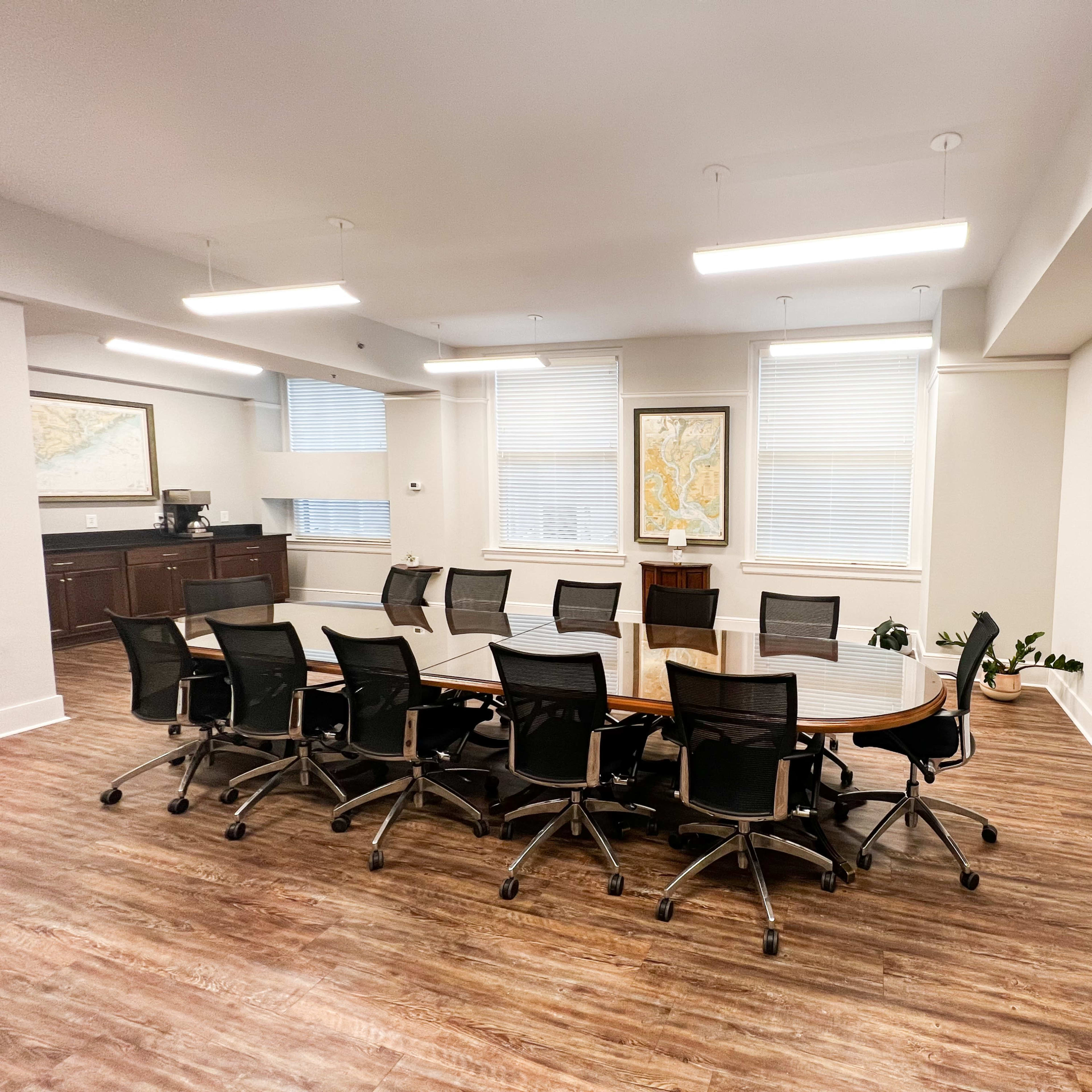 A modern conference room features a long wooden table surrounded by black office chairs, with large windows and a mounted television on the wall.