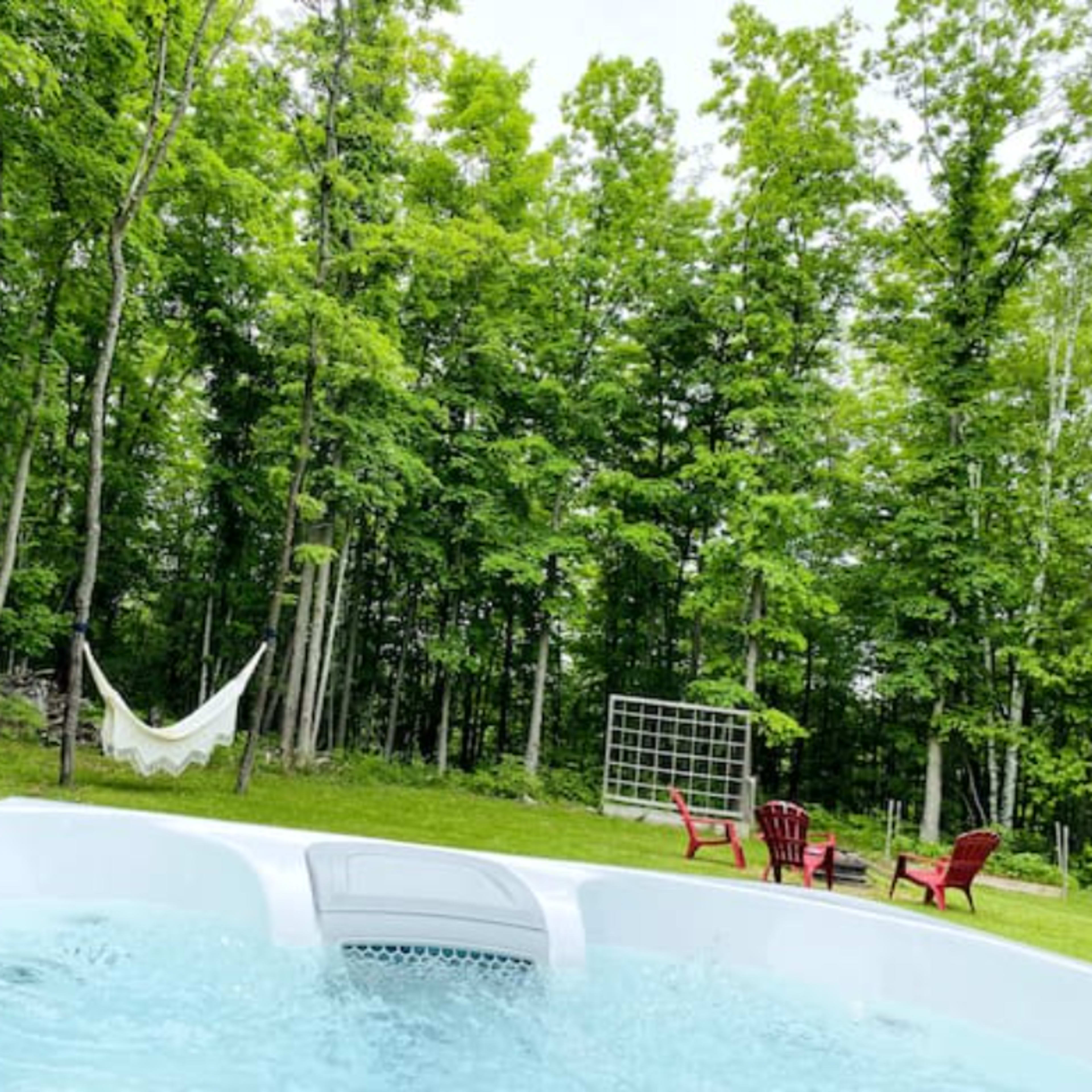 A hot tub is situated in a forested area, surrounded by green trees, with a hammock and red chairs nearby.