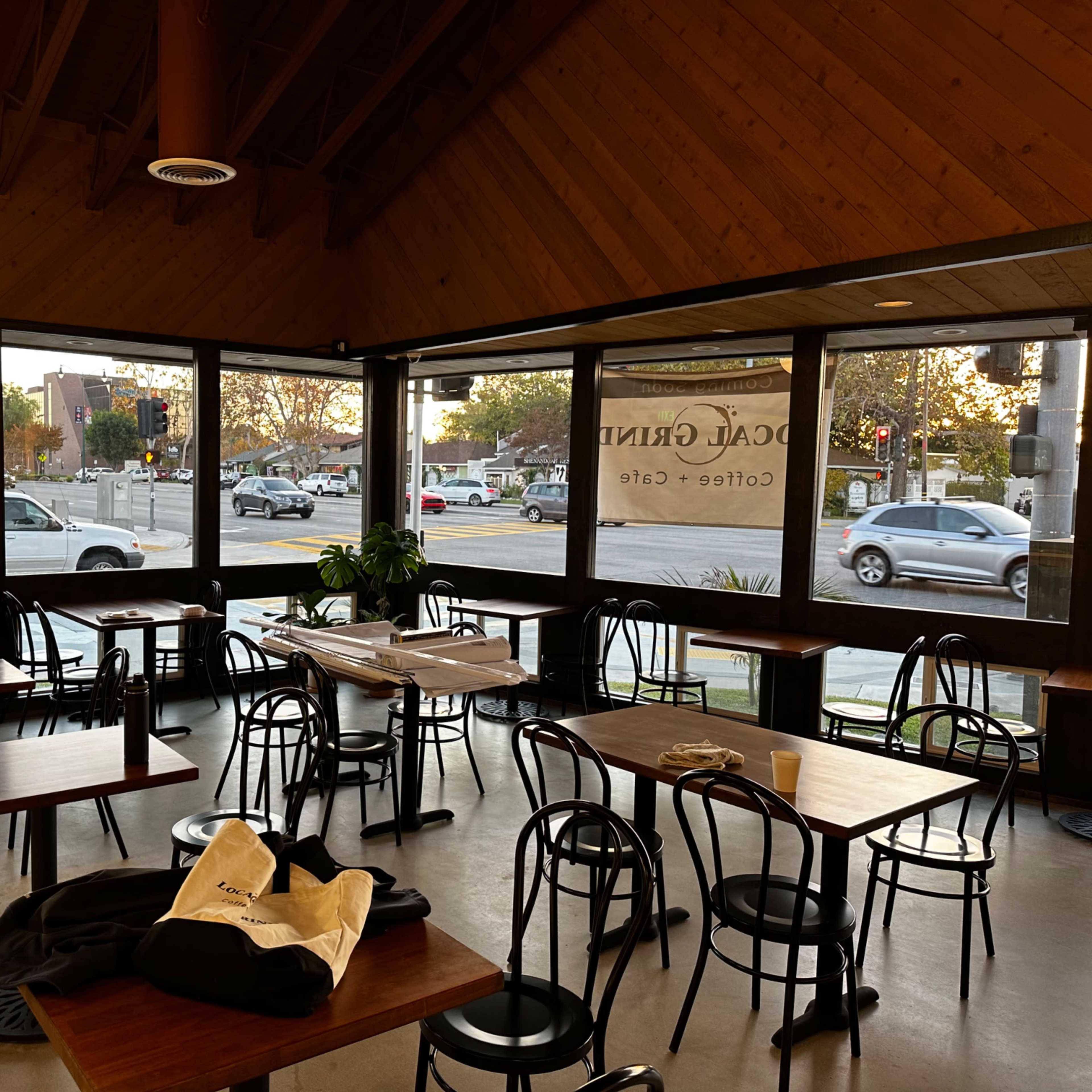 An interior of a café with wooden ceilings, several tables and chairs, and large windows facing a street.