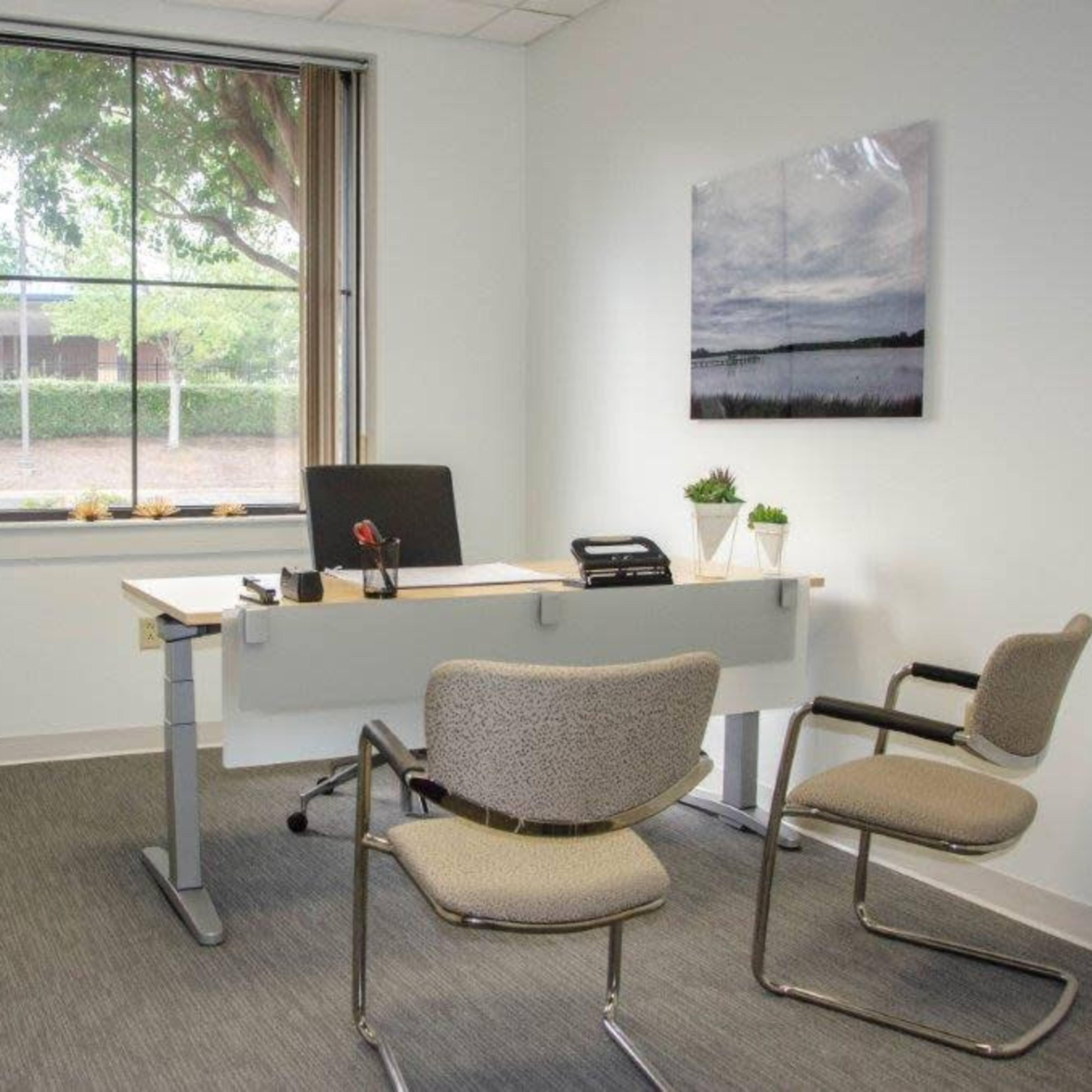 A simple office space featuring a desk with a chair, two additional chairs, a filing cabinet, and a window overlooking greenery.
