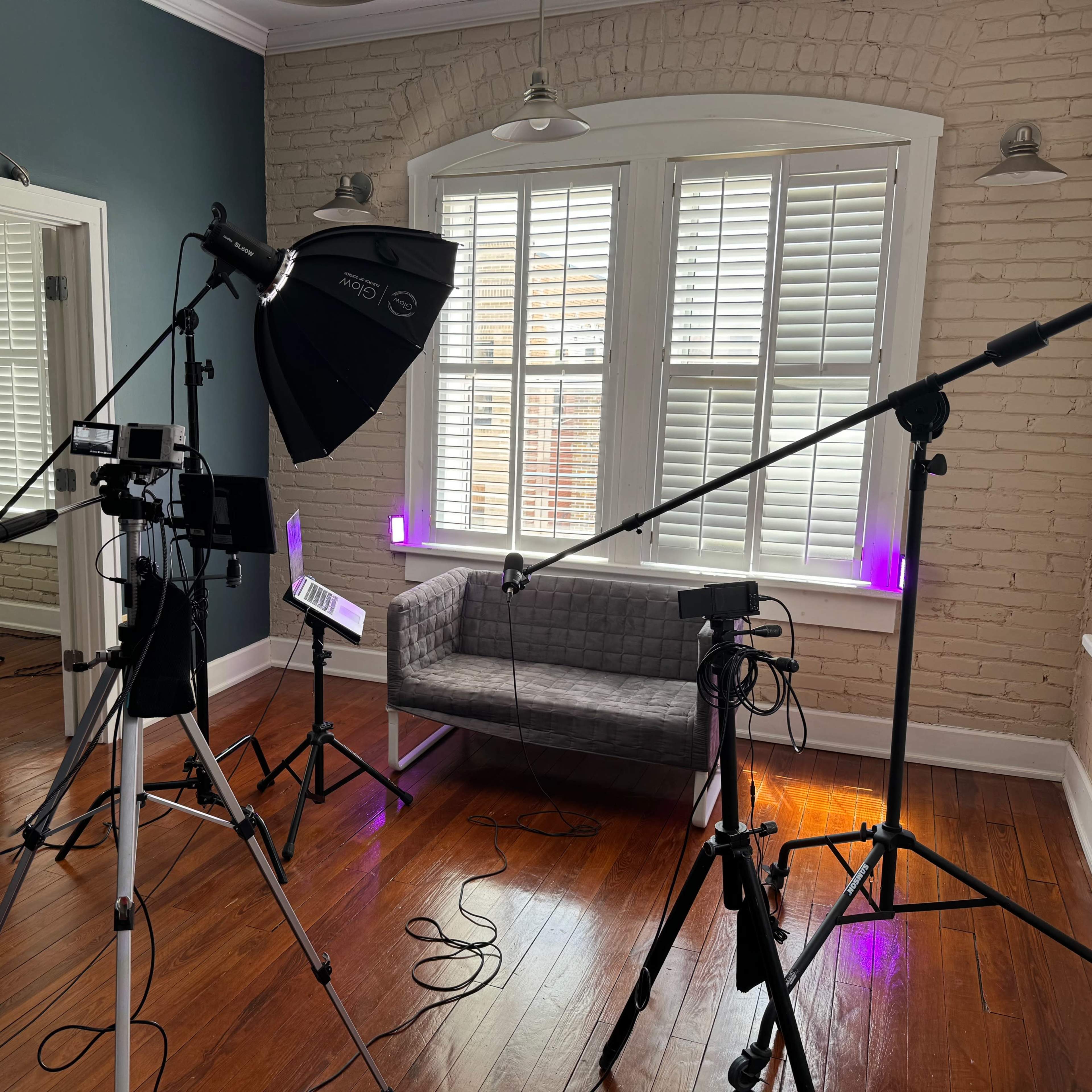 A photography studio setup with multiple tripods, lighting equipment, and a gray couch against a brick wall.