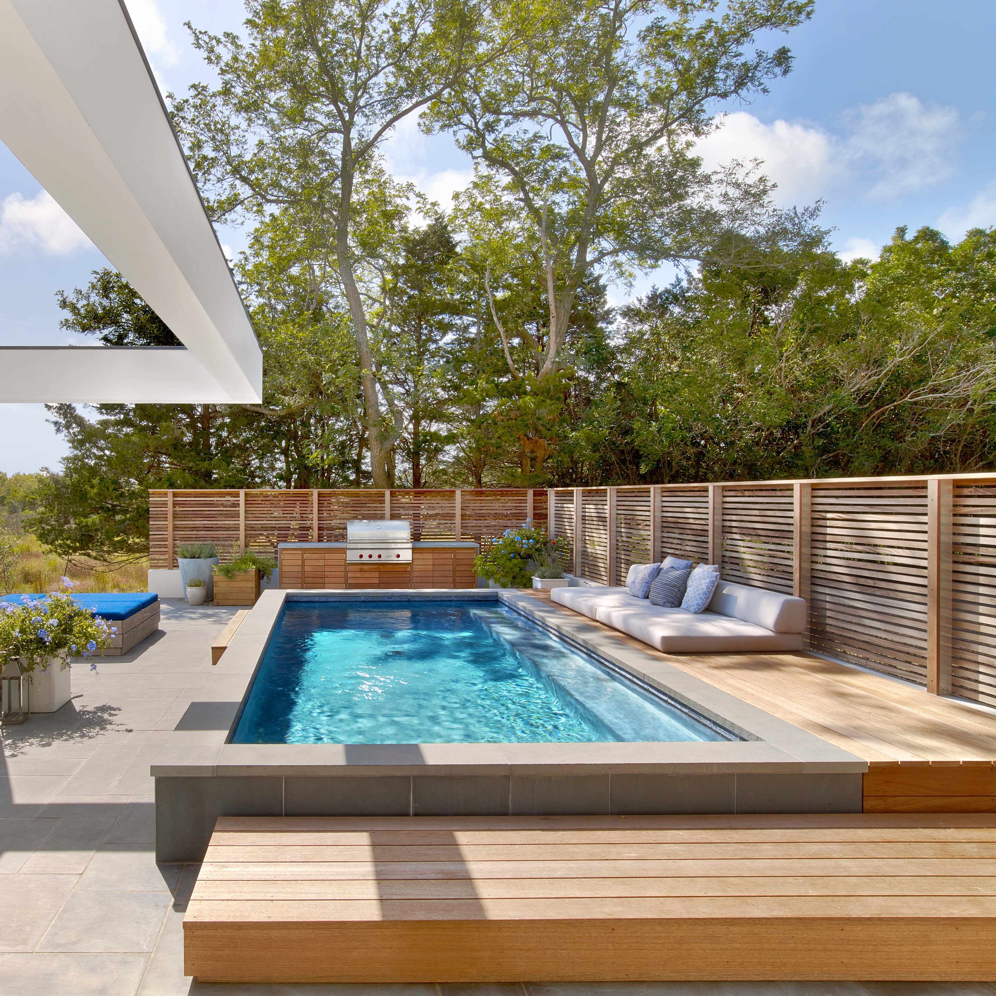 A modern outdoor space features a rectangular pool surrounded by a wooden deck, outdoor seating, and a barbecue area, set against a backdrop of trees.