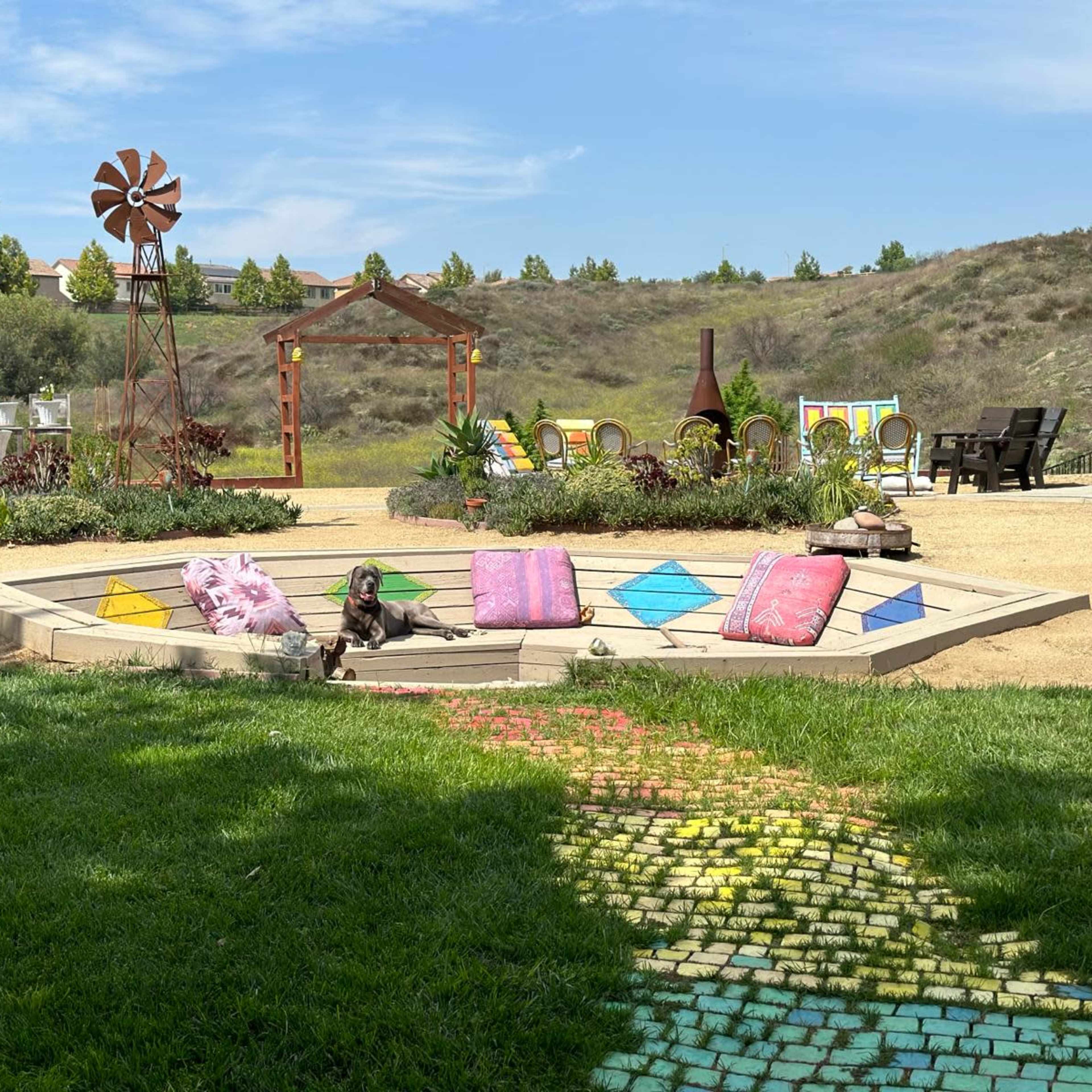 A landscaped backyard features a colorful pathway leading to a hexagonal seating area with cushions, surrounded by garden elements and a distant view of rolling hills.