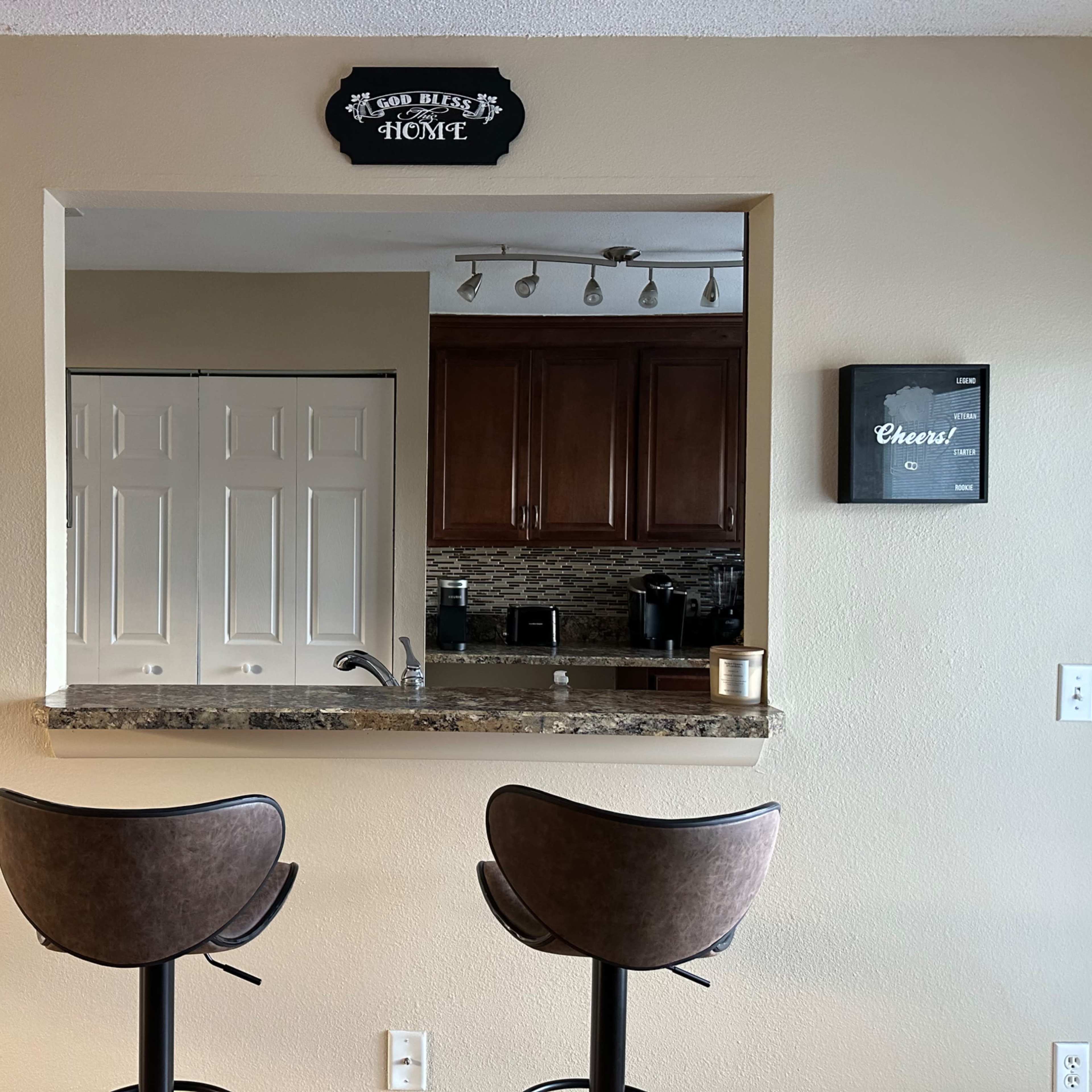 A bar area features two stools in front of a granite countertop with a wall-mounted sign above and cabinets in the background.
