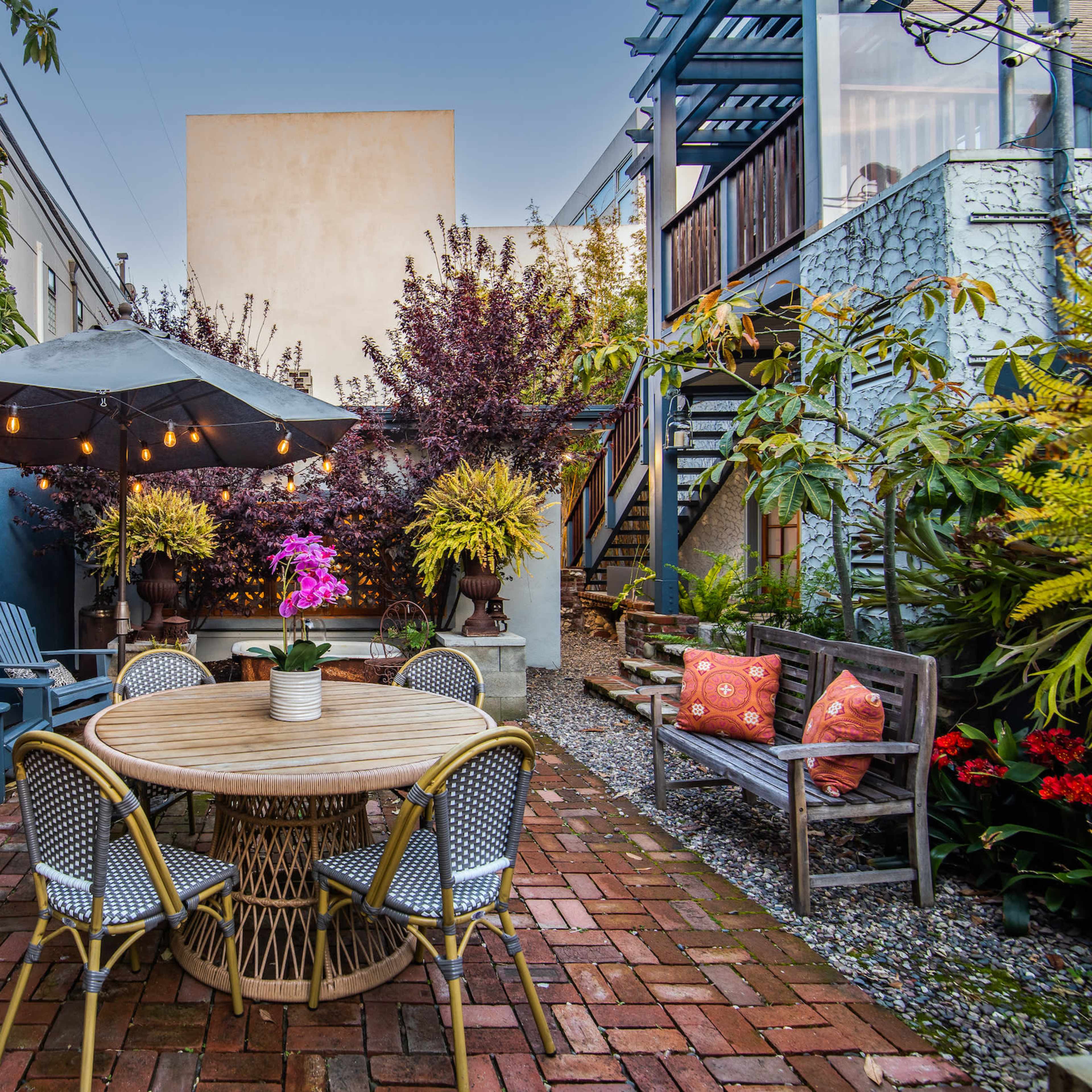 A garden patio with a round table surrounded by chairs, an umbrella, and various plants along the walls.