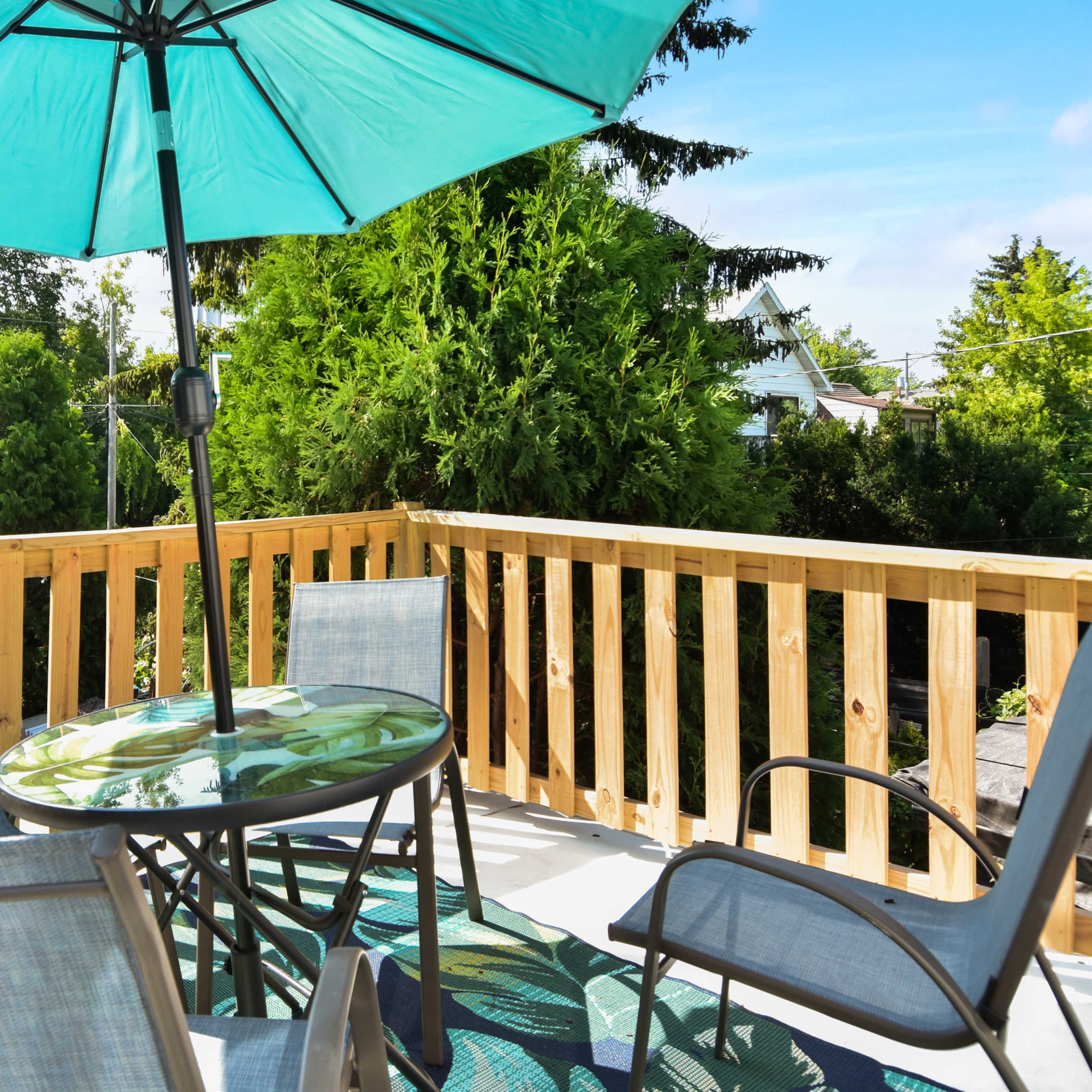 A small outdoor deck features a round table with a glass top, surrounded by four chairs and an umbrella, with greenery and distant houses visible in the background.